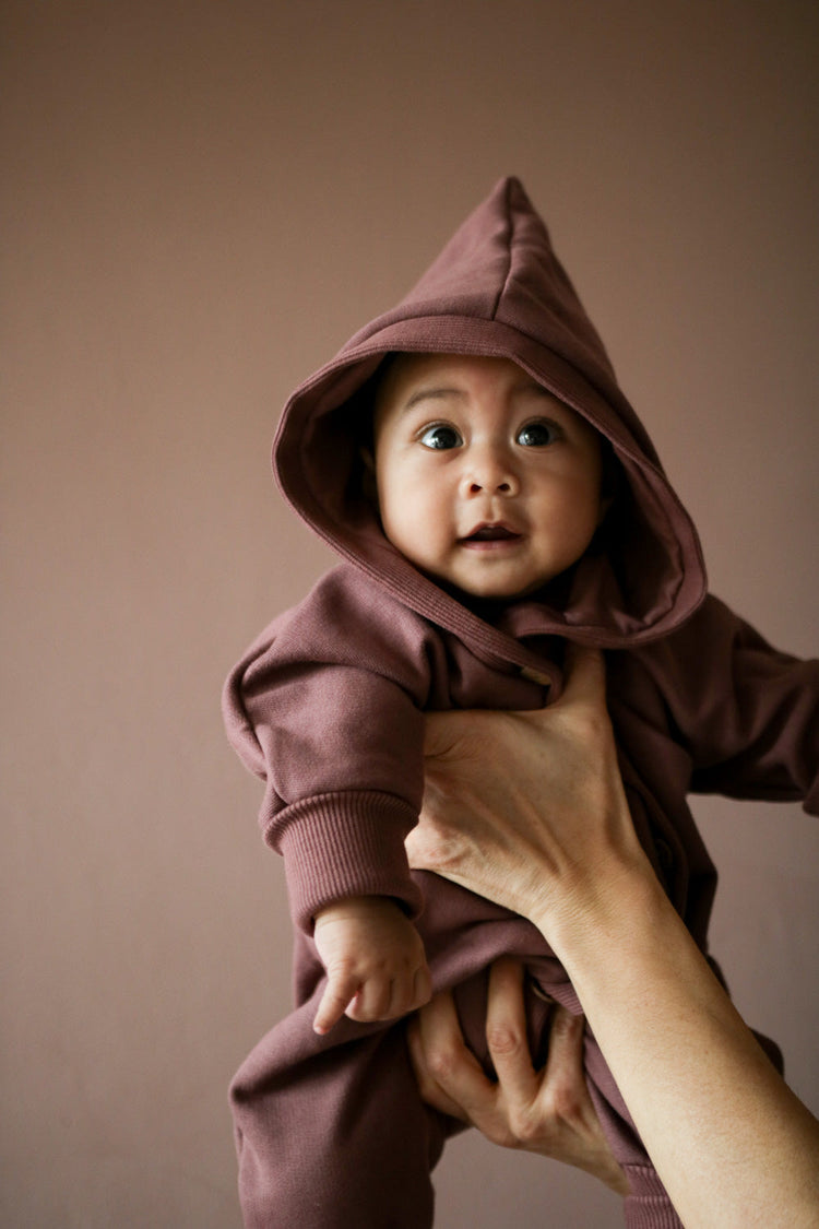 Baby mit Overall aus Bio-Baumwolle in lilabraun , gehalten von einem Erwachsenen.
