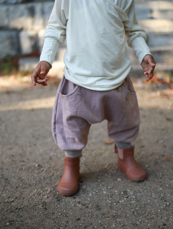 Ein Kind trägt ein Wickelshirt, Knickerbocker Leinen und braune Stiefel, stehend auf dem Boden im Freien. 