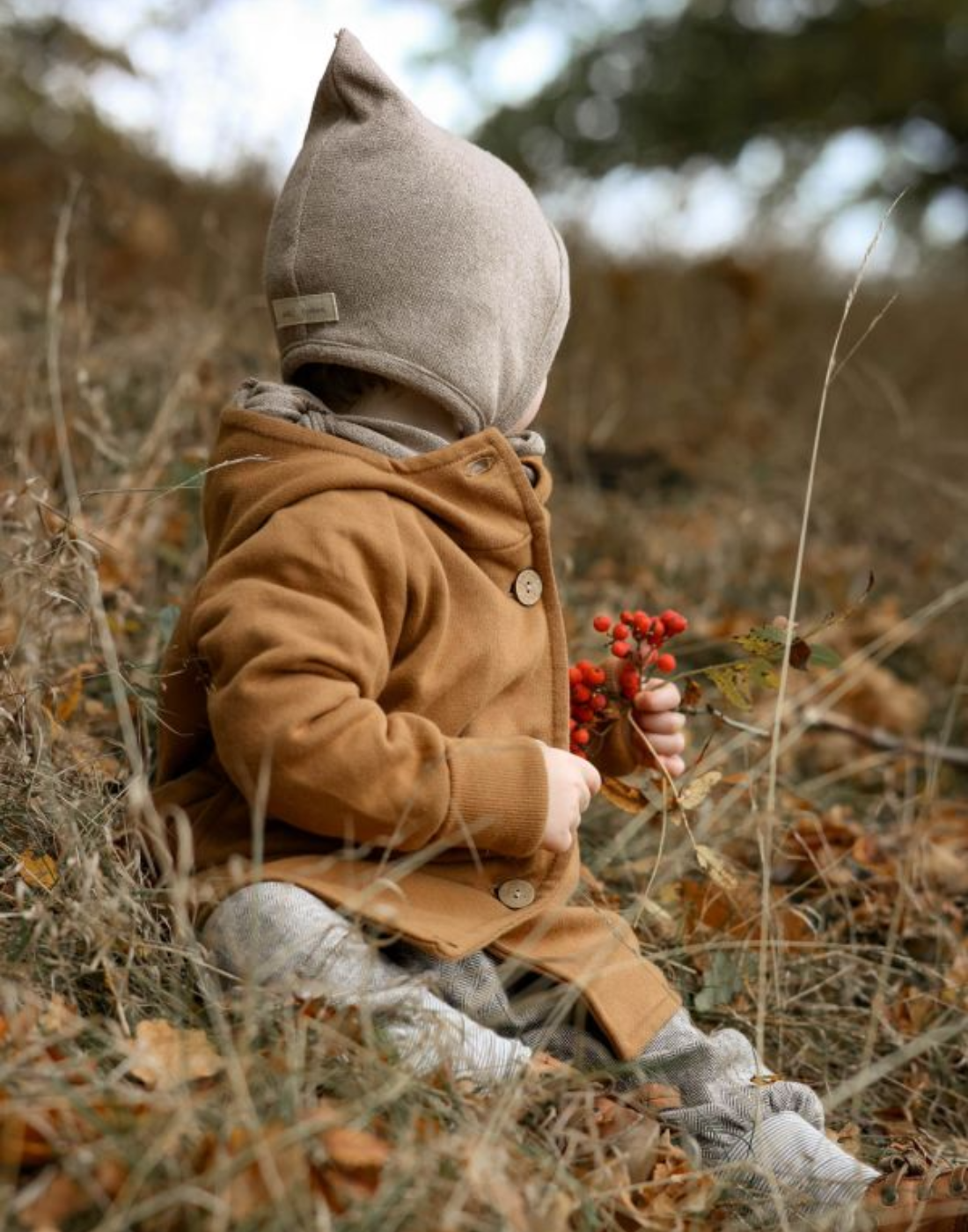 Ein Baby sitzt im Gras und hält einen roten Zweig. Es trägt eine Wichteljacke von Petit Cochon und befindet sich in einer natürlichen Umgebung.