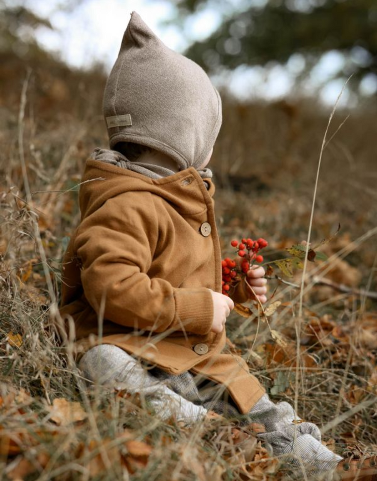 Ein Baby sitzt im Gras und hält einen roten Zweig. Es trägt eine Wichteljacke von Petit Cochon und befindet sich in einer natürlichen Umgebung.