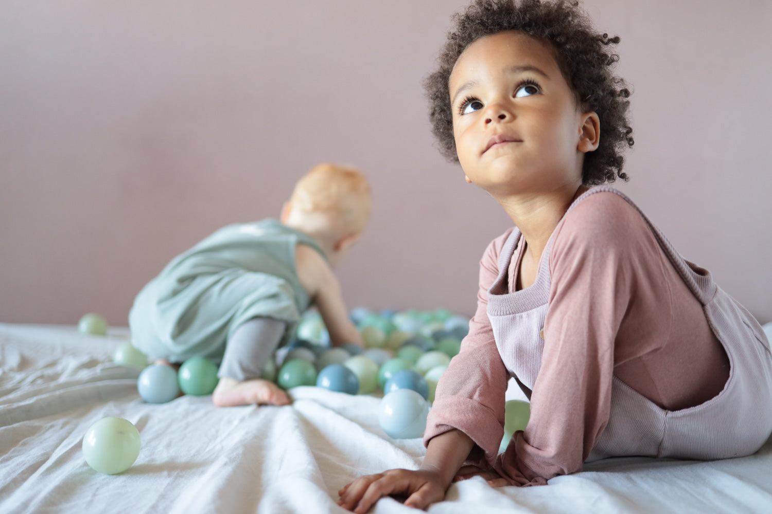 Eine Gruppe von Kindern spielt mit Bällen, während ein Baby auf einer Decke mit Bällen krabbelt. Nachhaltige Kinderkleidung, in Berlin genäht.