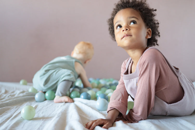 Eine Gruppe von Kindern spielt mit Bällen, während ein Baby auf einer Decke mit Bällen krabbelt. Nachhaltige Kinderkleidung, in Berlin genäht.