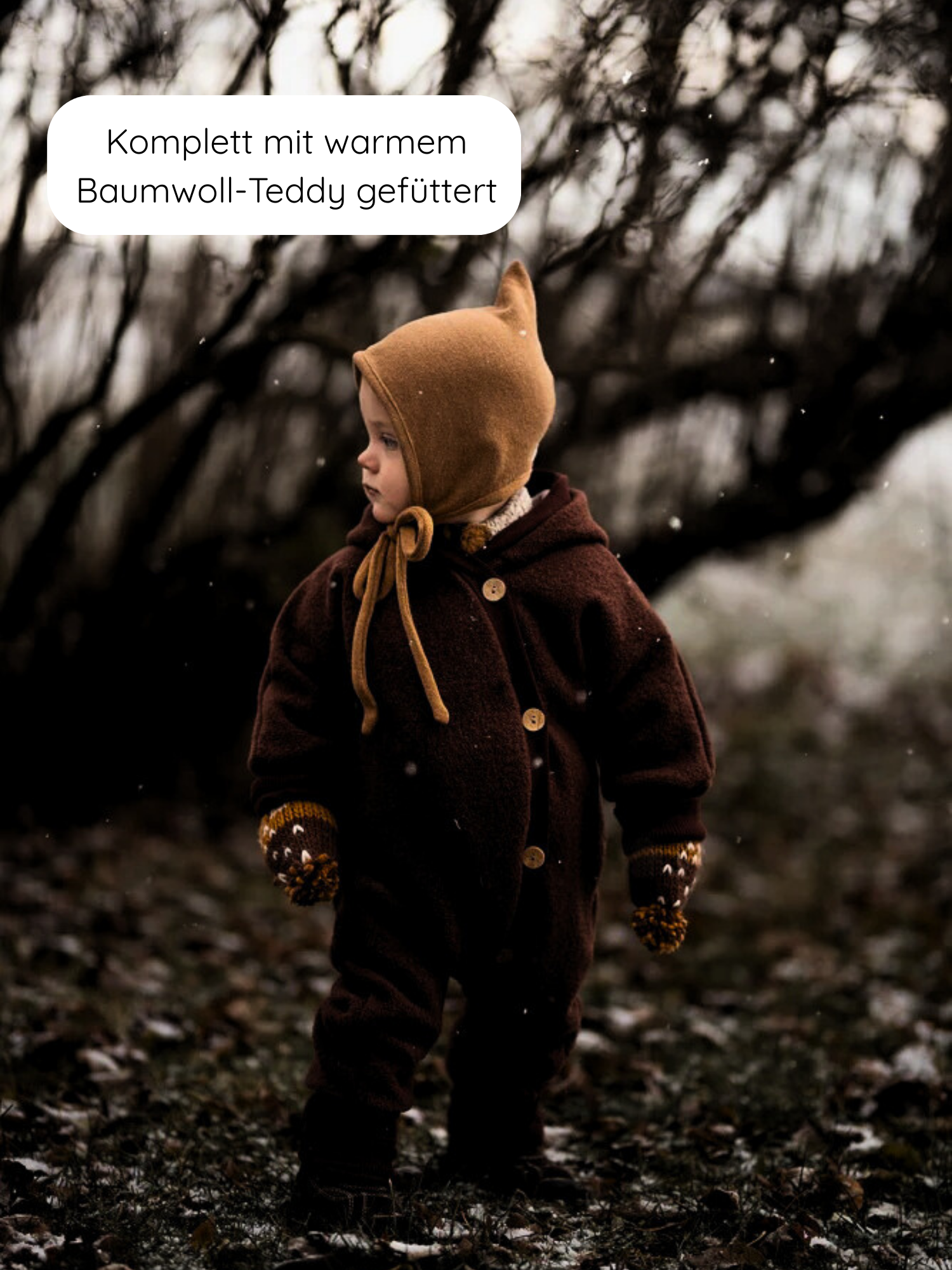 Kleinkind im Wald trägt Winter Wollwalk Overall mit Teddyfutter - Schoko, mit Holzknöpfen, passendem senfgelbem Mützchen mit Ohren und Handschuhen. Ideal für kalte Wintertage, hält warm und schützt vor Nässe.