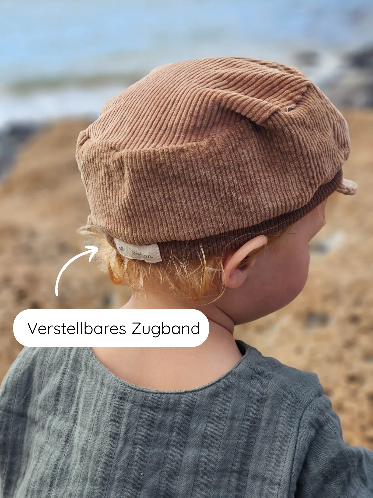 Kleinkind am Strand trägt die Michelmütze Breitcord Stretch - Weizen, einen warmen braunen Cordhut mit verstellbarem Zugband, ideal für kühle Tage, handgefertigt in Berlin.