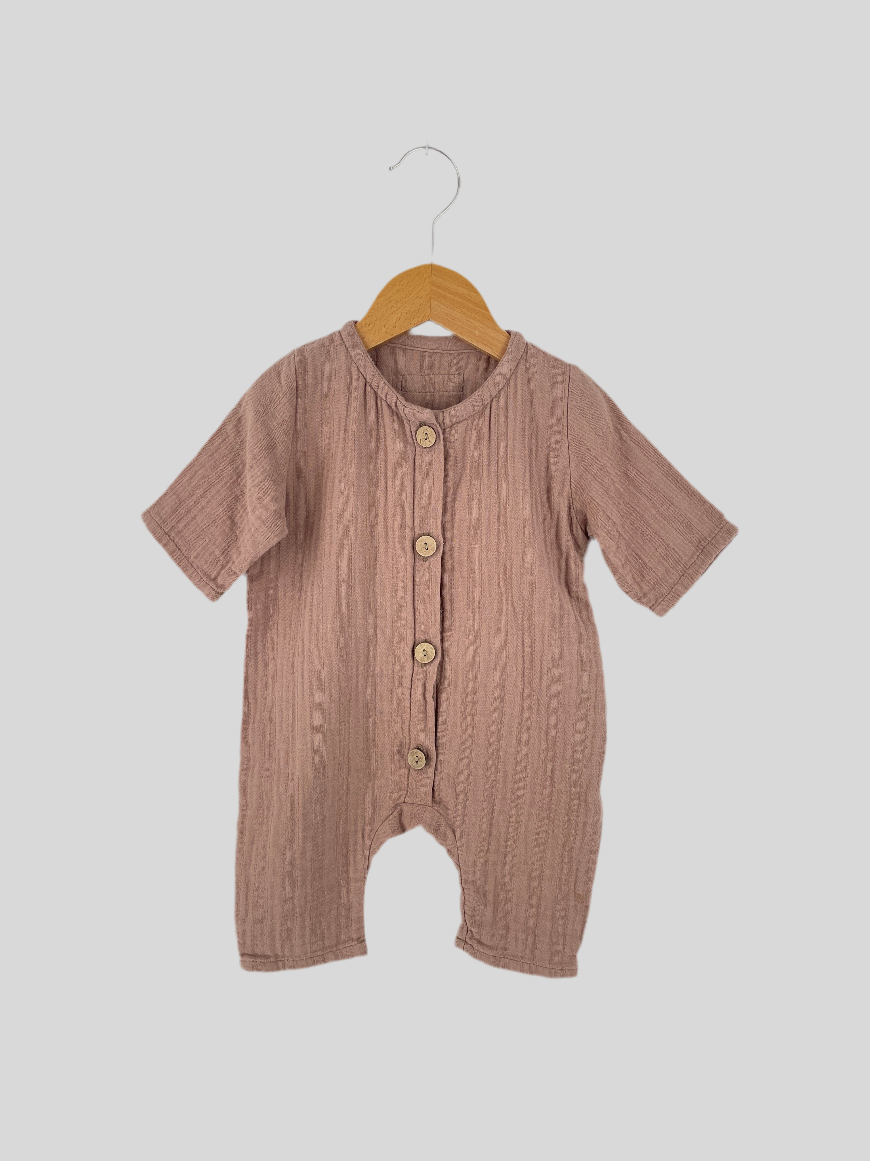 Ein Babystrampler Kopie Mini Overall Musselin Puder Gr. 0M aus weichem, leicht geknittertem Musselin-Stoff mit kurzen Ärmeln und Beinen, knopfbar, an einem Holzkleiderbügel vor neutralem Hintergrund hängend. Zustand: Sehr gut.