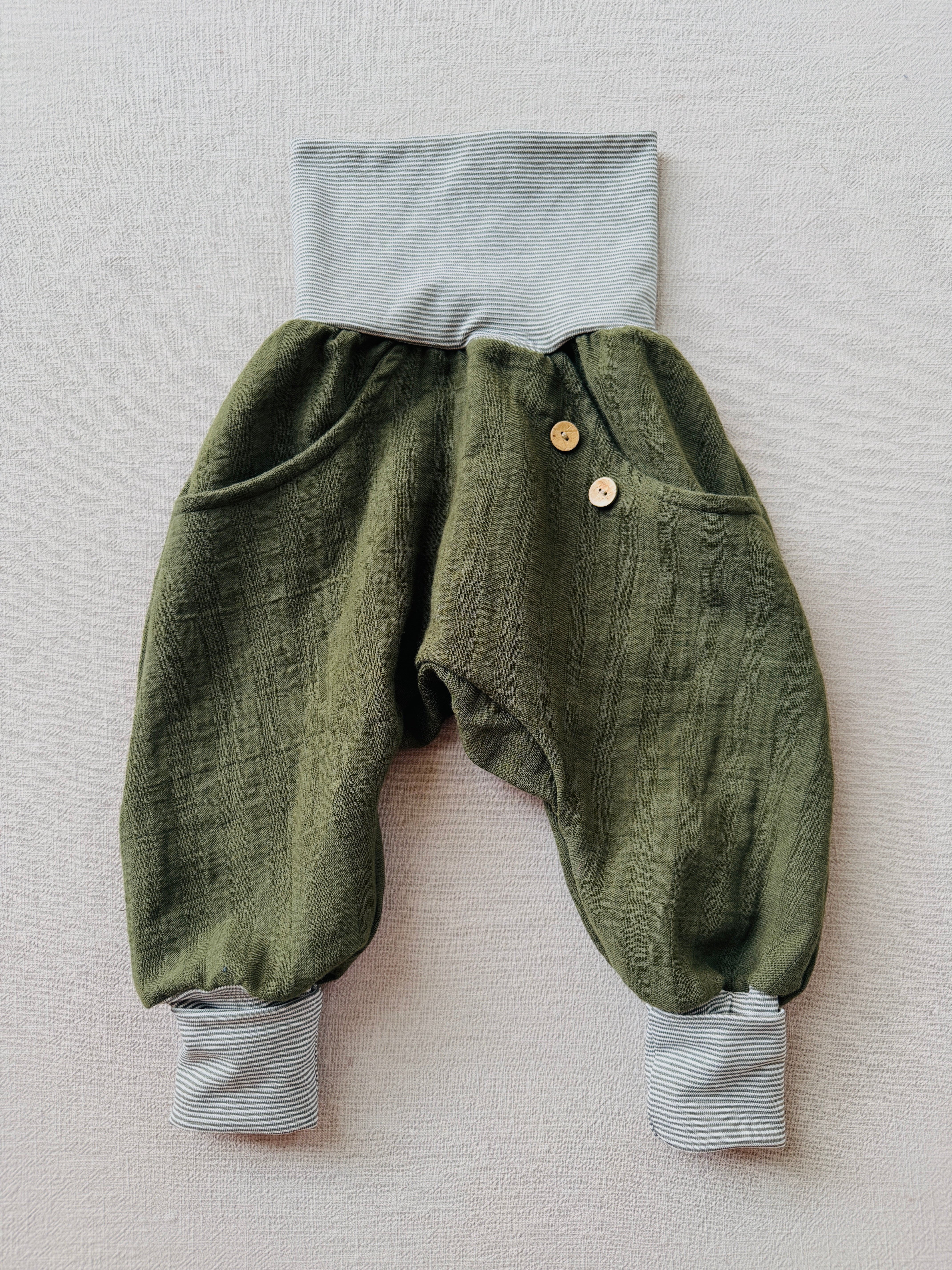 Knickerbocker Musselin aus 100% Bio-Baumwolle - Avocado: Olivgrüne, slouchige Kinderhose mit grauen Streifenbündchen, Seitentaschen und zwei Holzknöpfen vorne, ideal für Bewegungsfreiheit und Komfort.