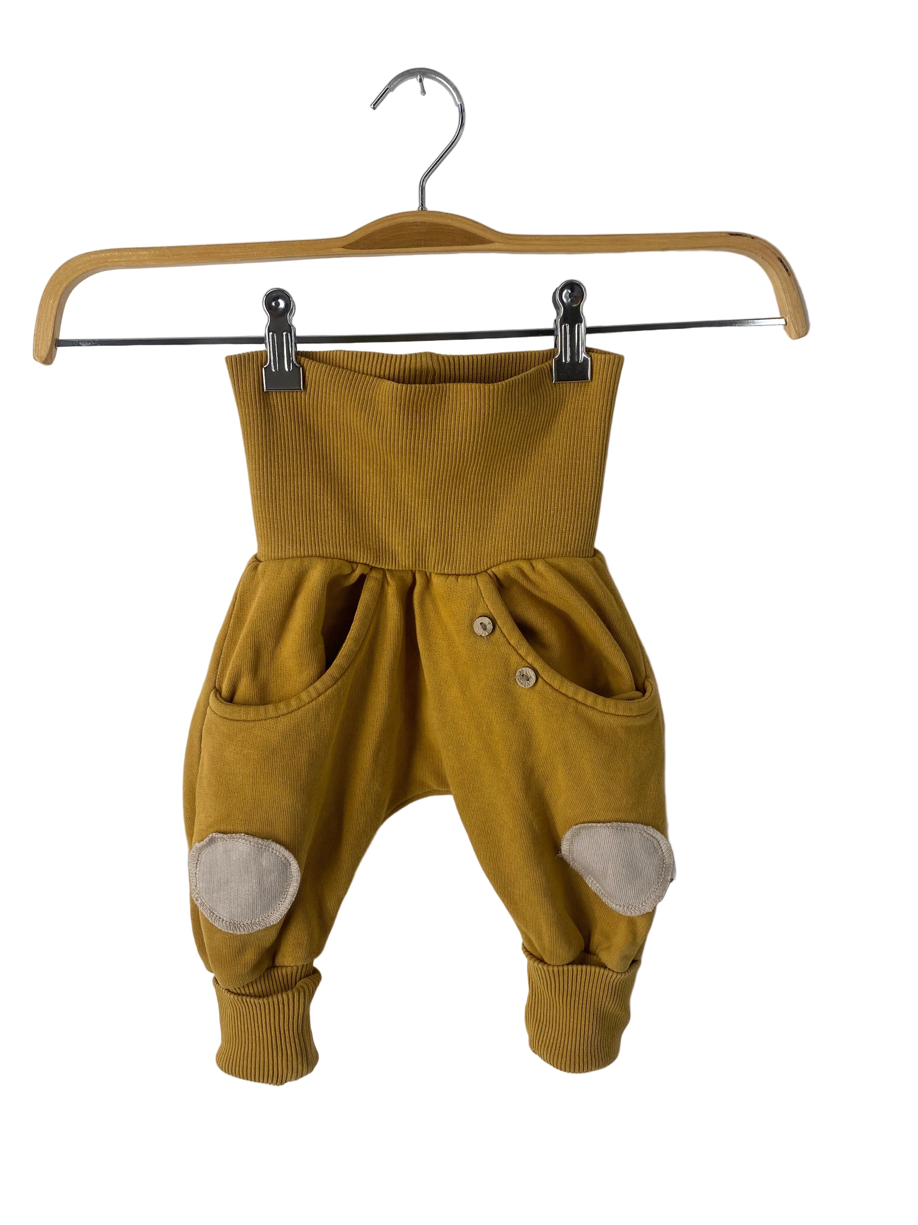 Knickerbocker Bio Sweat Kurkuma Gr 6M: Senfgelbe Babyhose mit tiefem Schritt, breitem Rippbund, Seitentaschen mit Holzknöpfen, beigen Knieflicken und Rippbündchen am Knöchel. Zustand: gut, zwei aufgenähte Flicken.