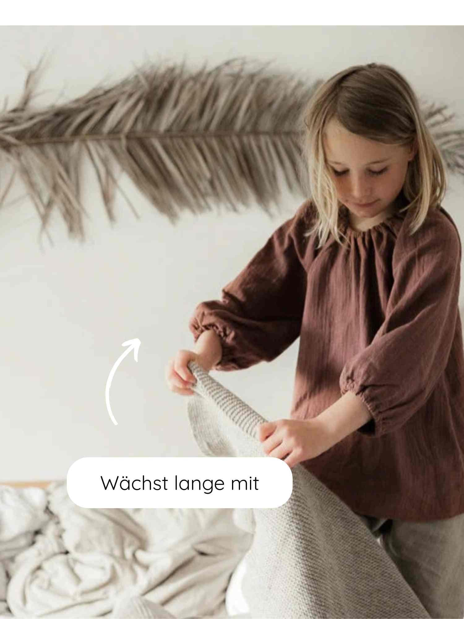 Kind trägt leichtes Blüschen Musselin - Malve, locker geschnitten und ideal für warme Tage. Es hält einen grauen Strickdecke, umgeben von neutralen Tönen und natürlicher Dekoration.