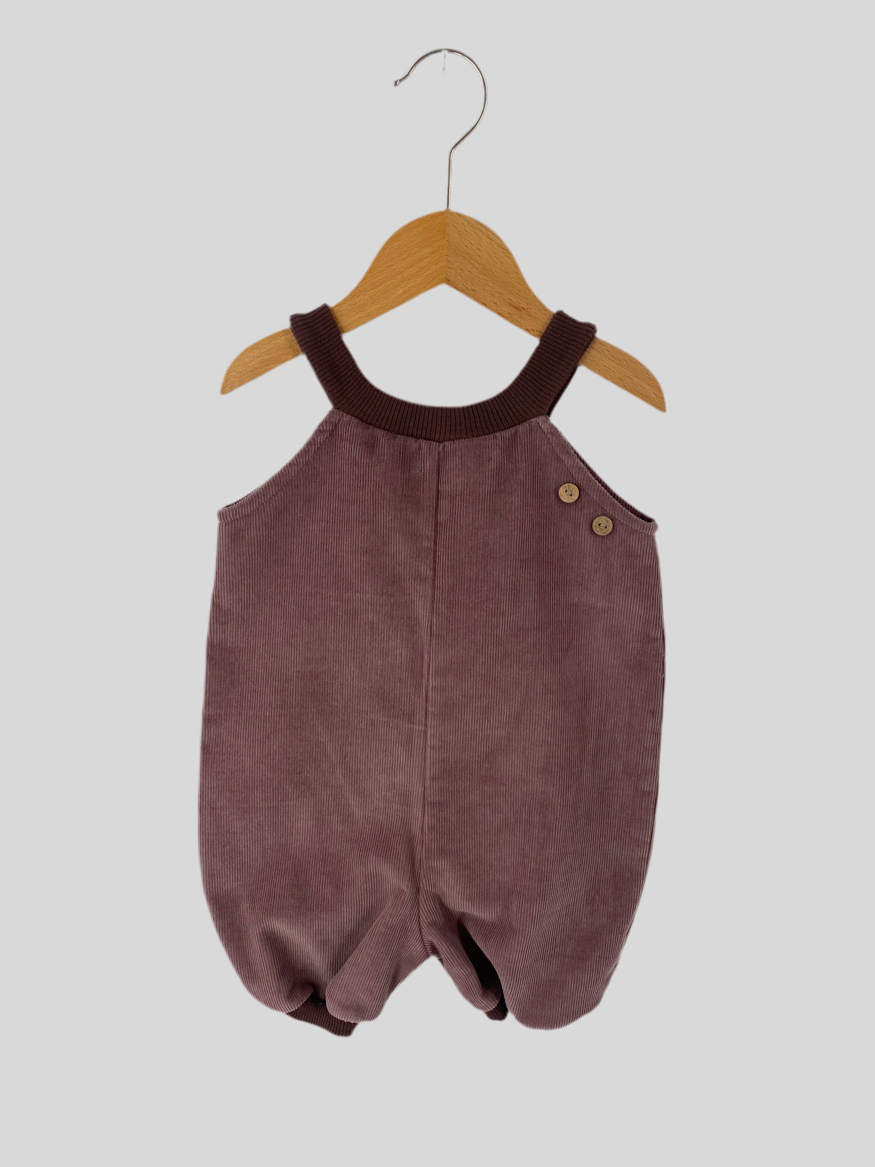 Kurze Baby Schlupflatzhose Feincord Malve Gr 3M: Ein ärmelloser Baby-Einteiler aus Feincord mit Strickträgern, zwei Holzknöpfen an der Seite und blusonartigen Beinöffnungen, auf einem Holzbügel hängend. Zustand: neu.