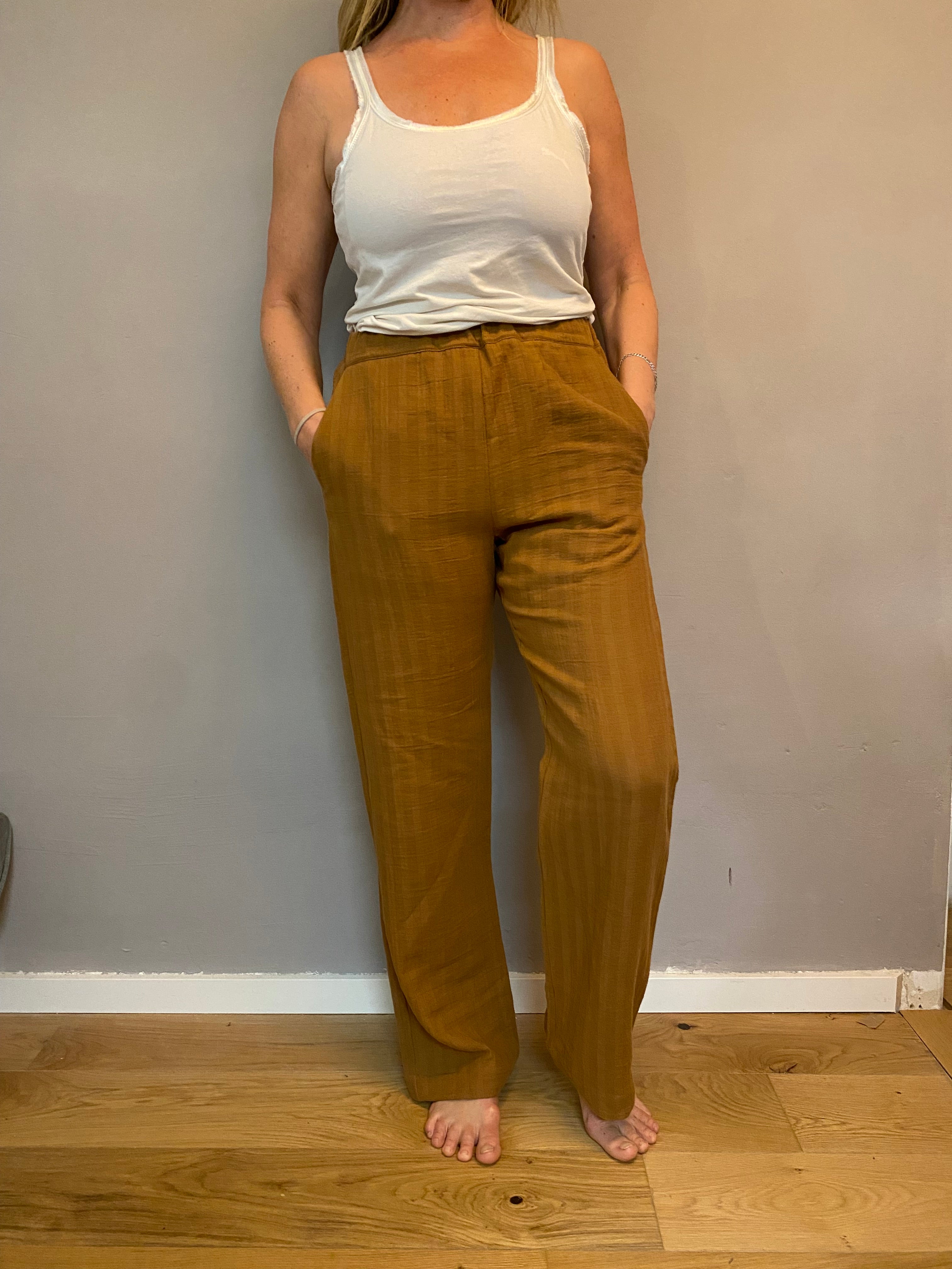 Person trägt Palazzo Hose Musselin Sahara Gr. M, steht barfuß auf Holzboden. Hose hat lockeren Sitz, Hände in Taschen. Sichtbar von Taille bis Füße, kombiniert mit weißem Tanktop.