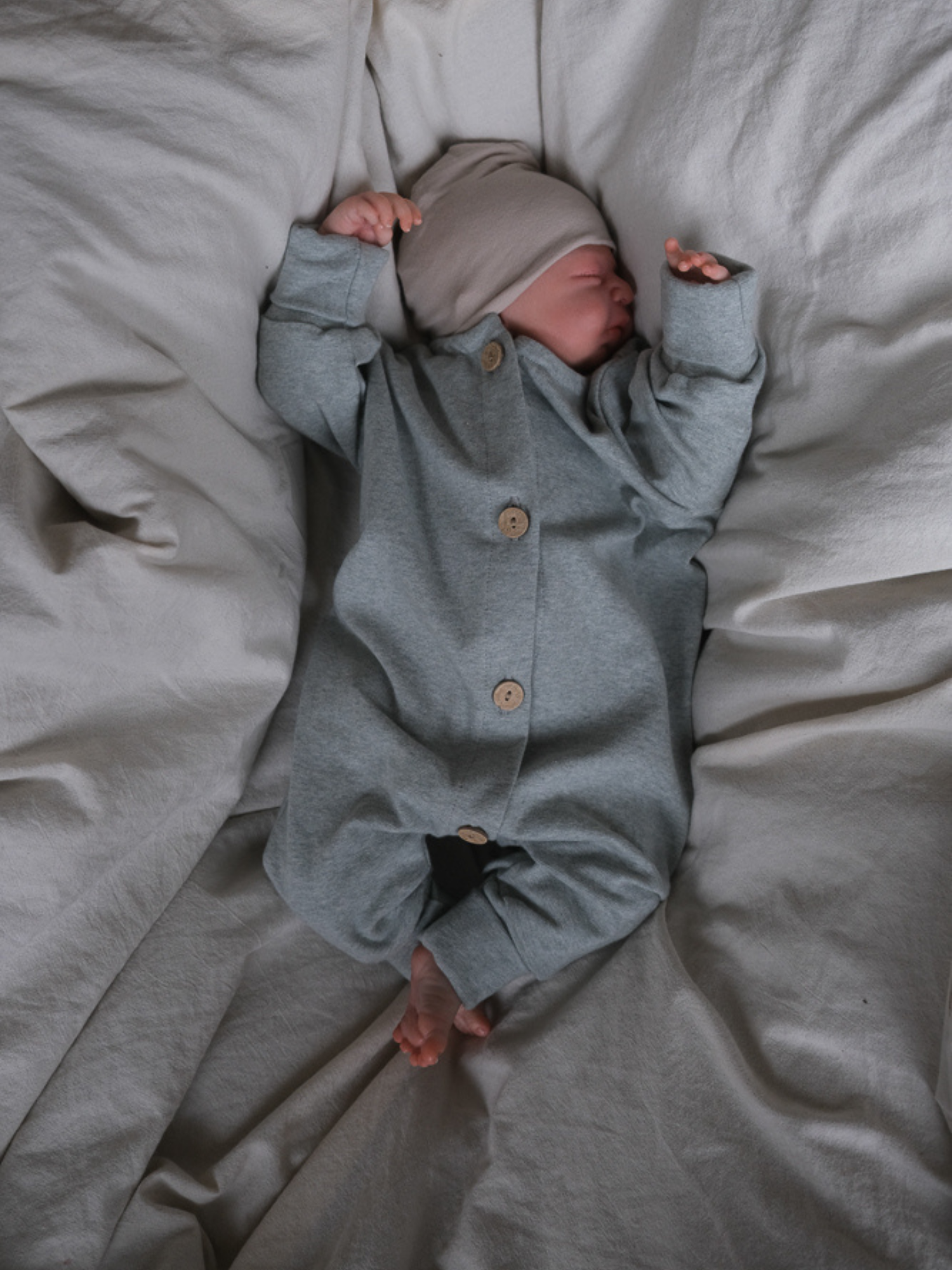 Neugeborenes in Mini Overall Feinstrick - Mint Meliert, mit Knopfleiste und weichem Strickstoff, schläft friedlich auf zerknitterter Bettwäsche, Arme über dem Kopf, Beine sanft gekreuzt.