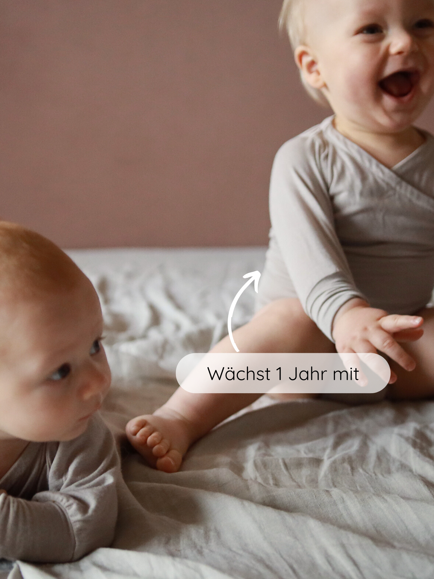 Zwei Babys in sandfarbenen Loopbody Kaschmir-Mix, eines sitzend lachend, das andere auf dem Bauch liegend. Ein Pfeil zeigt auf das sitzende Baby mit dem Text „Wächst 1 Jahr mit“.