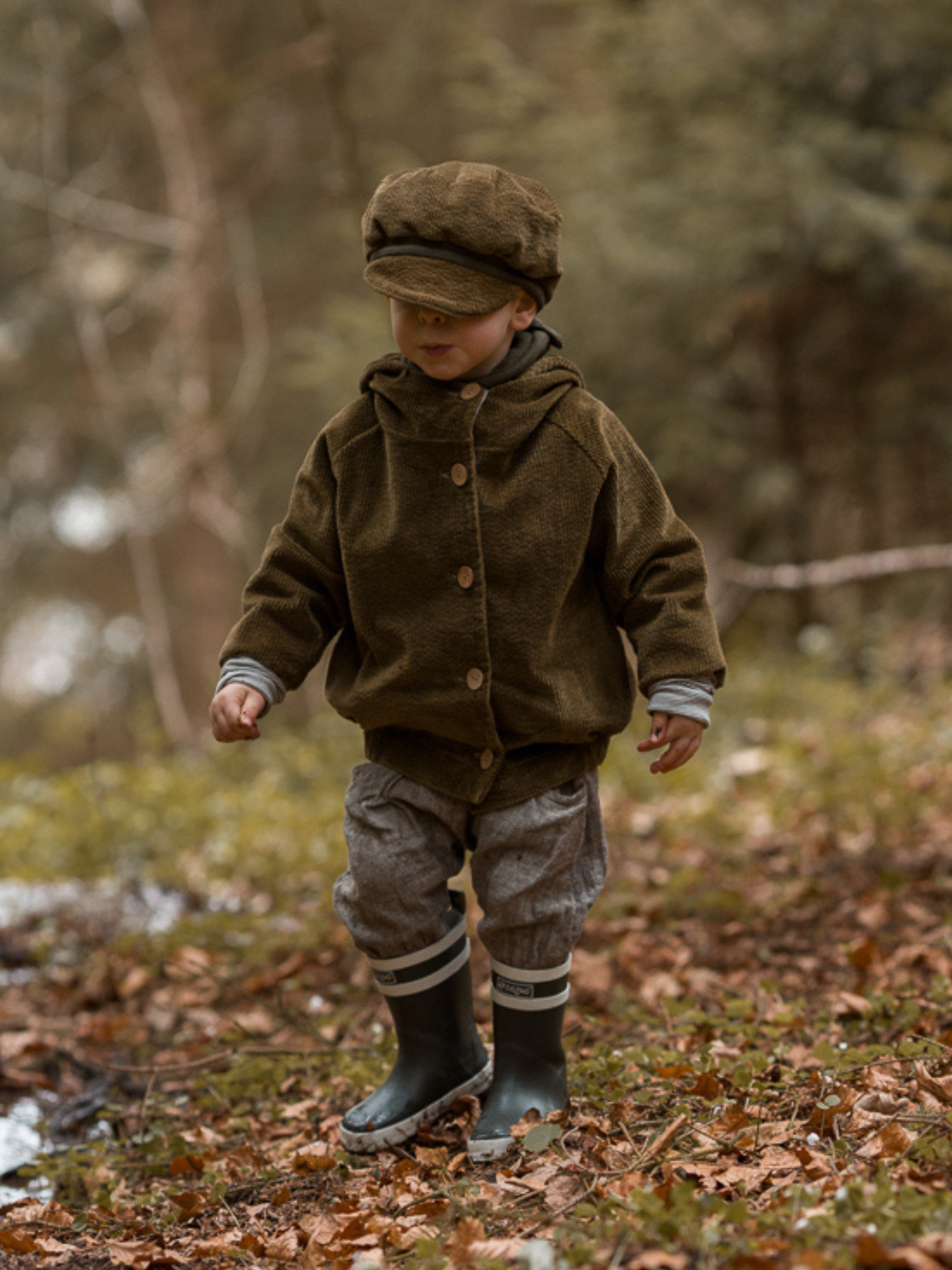 Ein Kleinkind trägt die Wichteljacke Breitcord Stretch - Kiwi, erkennbar an den krempelbaren Ringel-Armbündchen und der Wichtelkapuze, während es vorsichtig durch eine mit Laub bedeckte Waldlandschaft spaziert.