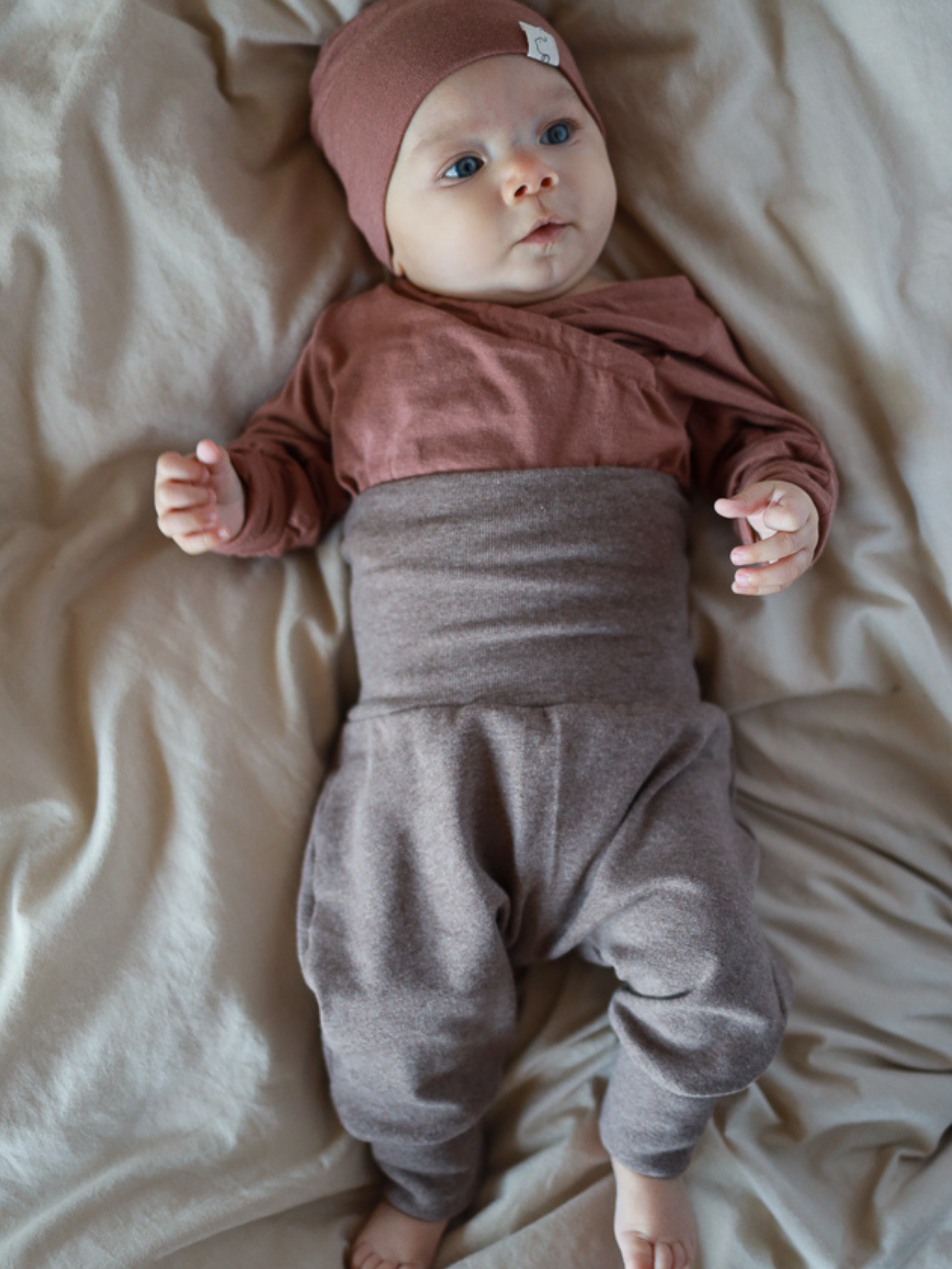 Babyknicker Feinstrick - Praline Meliert: Ein Säugling trägt eine rostbraune Mütze und ein Oberteil, kombiniert mit graubraunen, locker sitzenden Babyknickern aus weichem, elastischem Feinstrick-Jersey, ideal für Bewegungsfreiheit.
