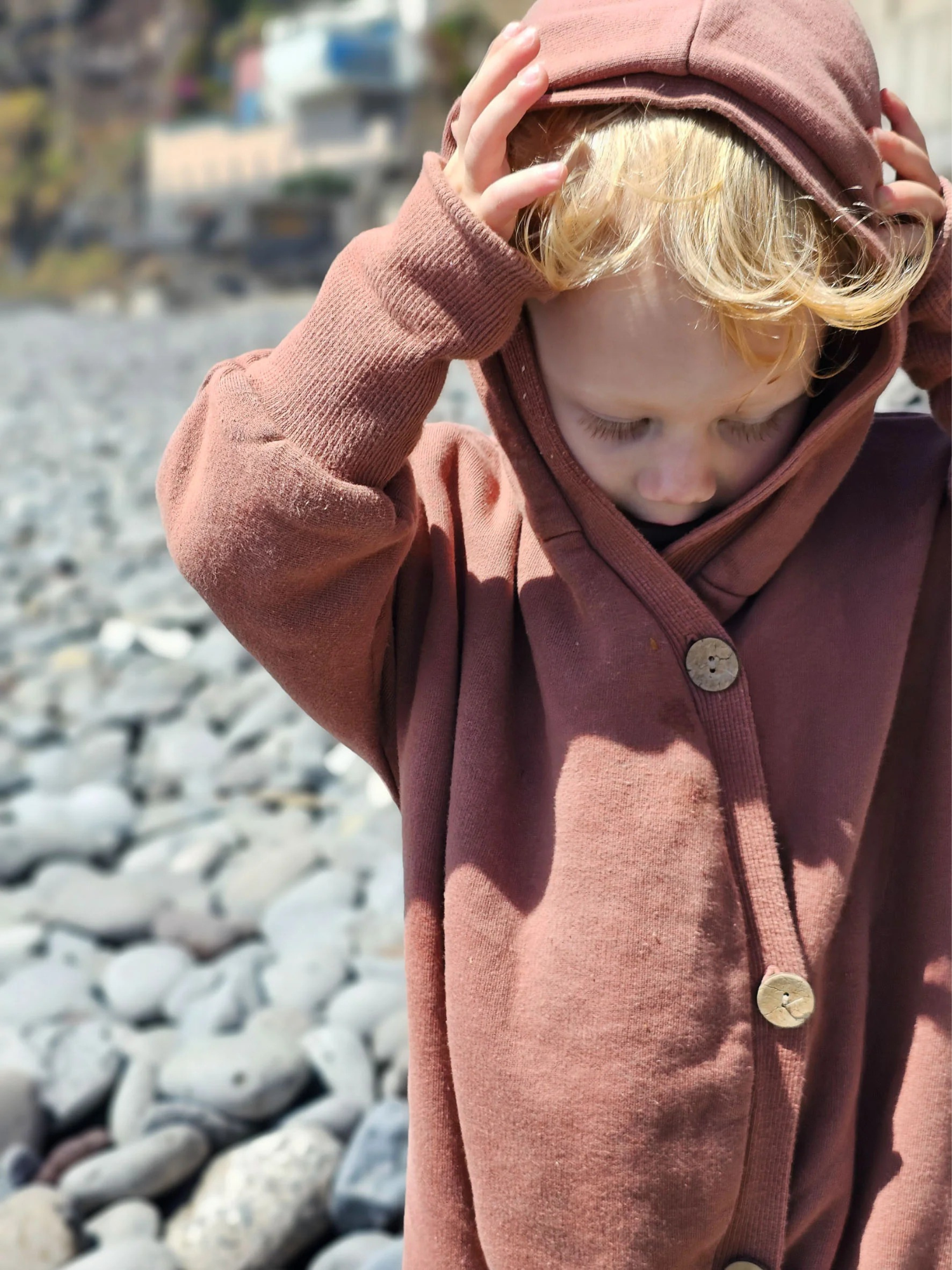Kleinkind im Kuschelanzug Bio-Sweat - Zimt, mit grob geschnittener Wichtelkapuze, zieht die Kapuze über den Kopf, stehend auf einem Kiesstrand.
