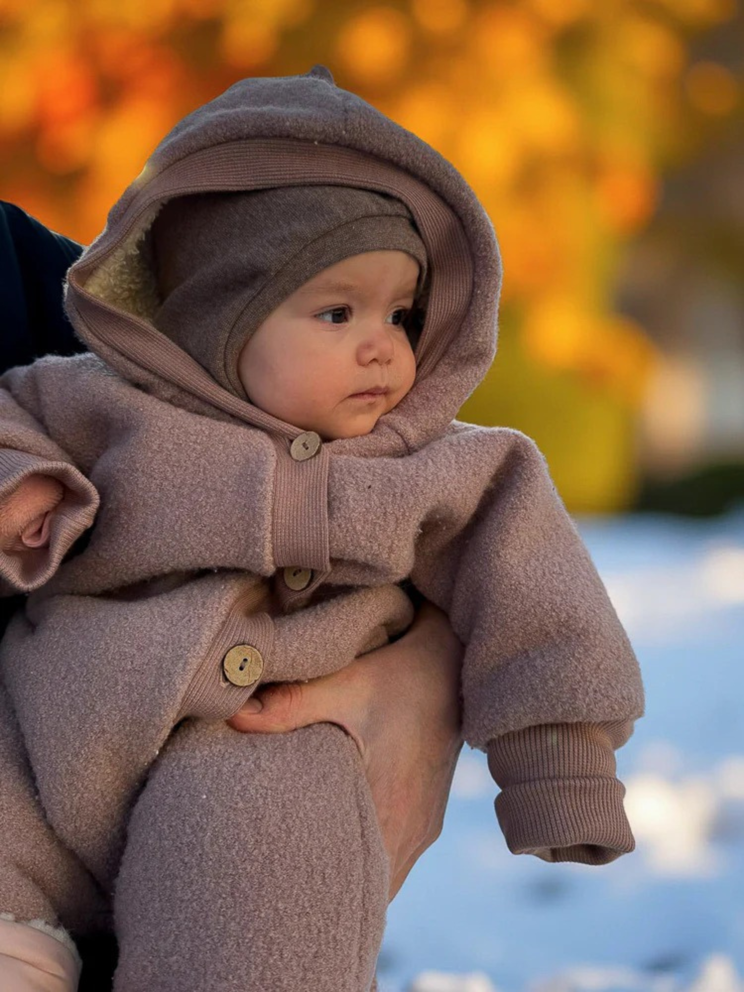 Ein Baby trägt die Schlupfmütze Feinstrick-Jersey - Hafer Meliert, die Kopf, Ohren und Hals schützt. Ein Erwachsener stützt sanft, während herbstliches Laub und Schnee den Hintergrund bilden.