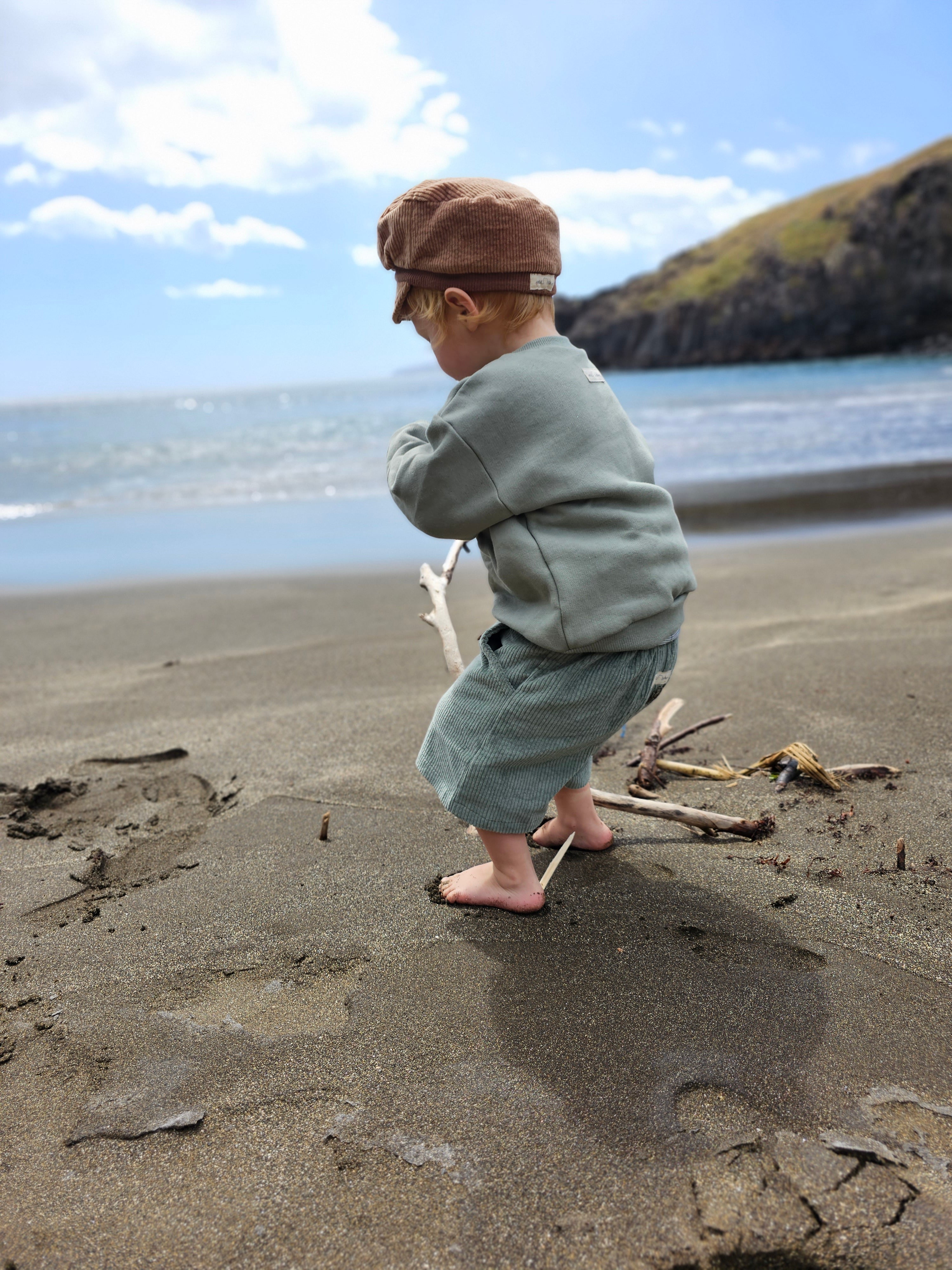 Kleinkind am Strand trägt eine Kurze Buxe Breitcord - Aqua, hält einen Stock im Sand. Die Hose ist robust, weich und ideal für Abenteuer.