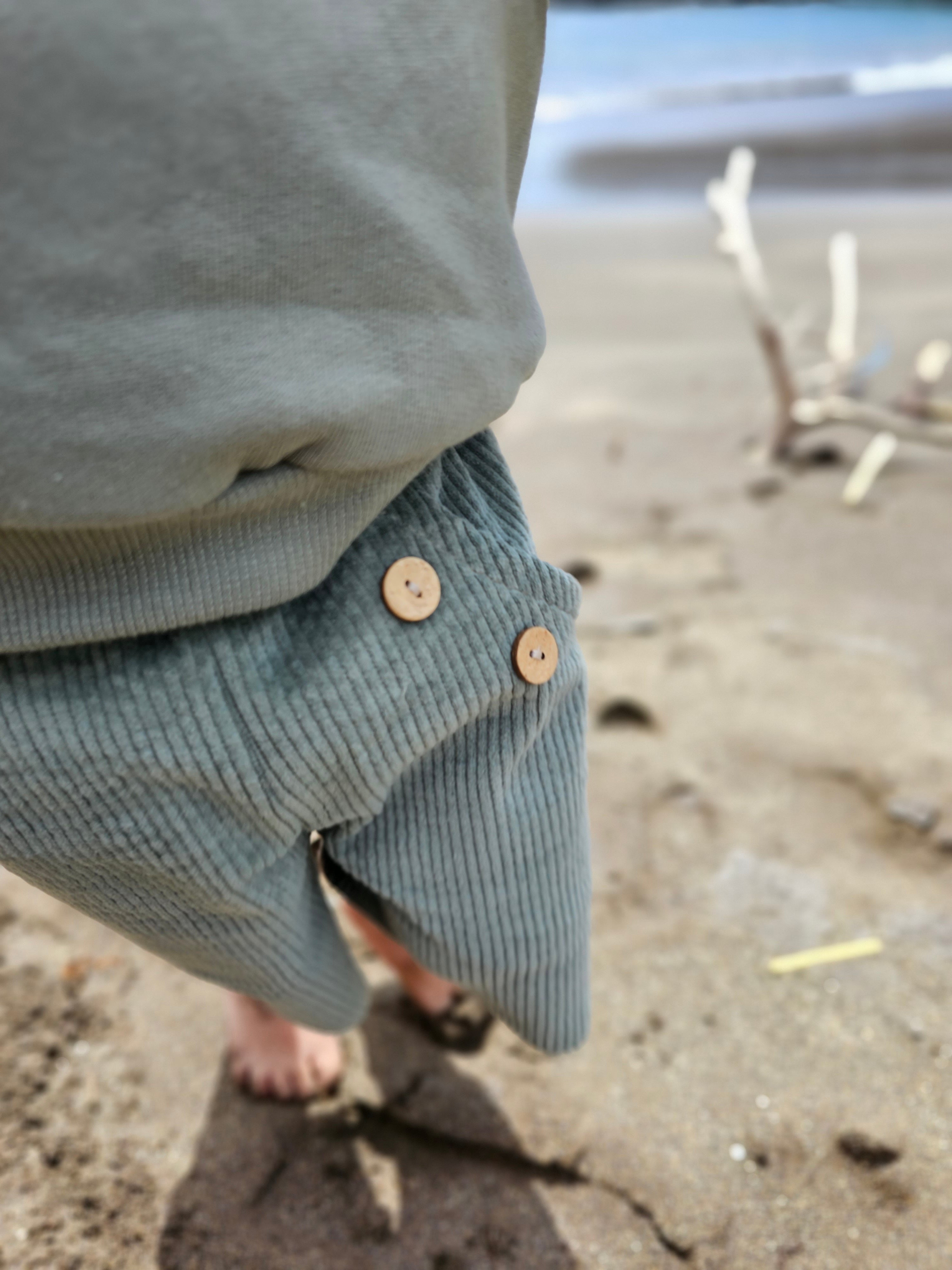 Ein Kleinkind läuft barfuß am Strand und trägt die Kurze Buxe Breitcord - Aqua, eine robuste, weiche Cordhose mit zwei Holzknöpfen am Bund, ideal für Abenteuer.