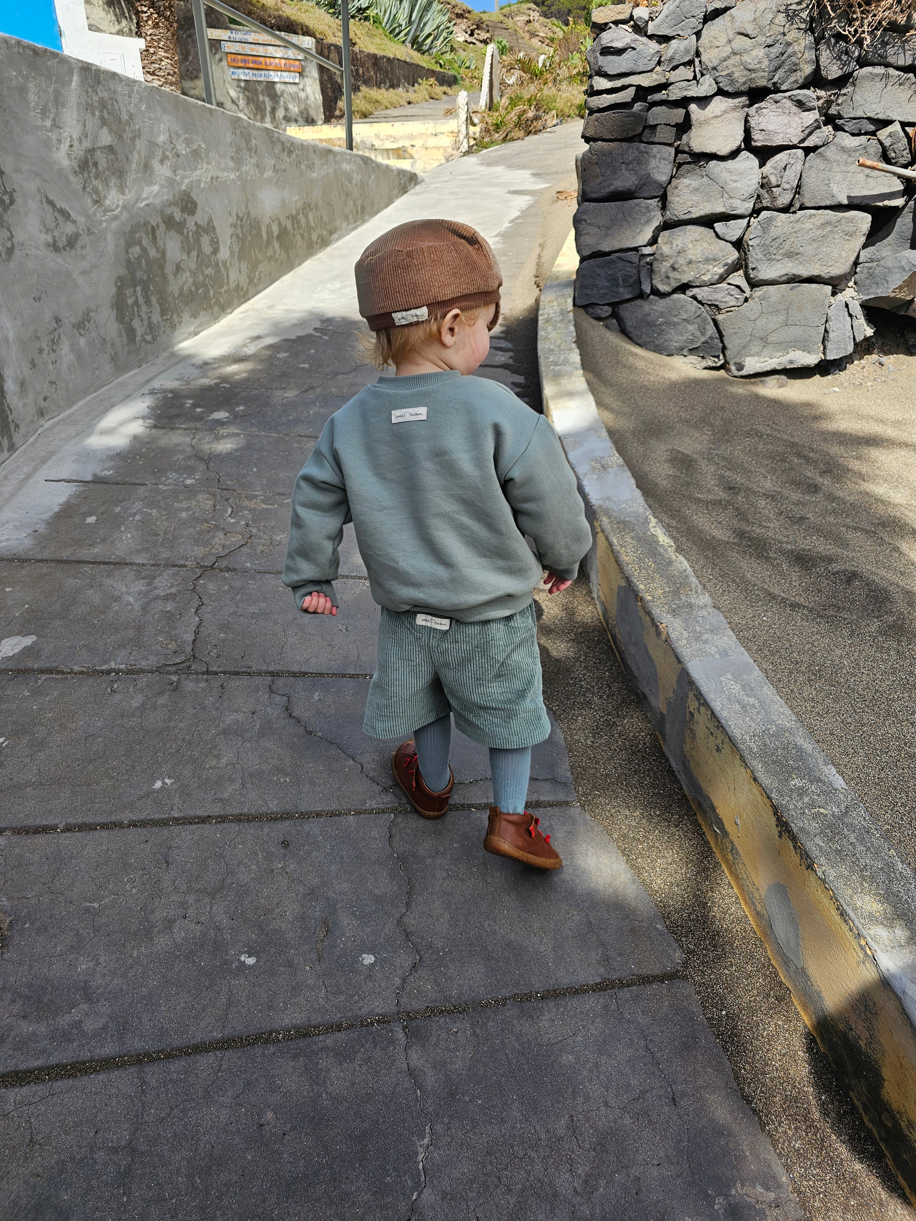 Kleinkind von hinten in Kurze Buxe Breitcord - Aqua, über blauen Strumpfhosen, auf Betonweg. Trägt braune Lederschuhe mit roten Schnürsenkeln und braune Strickmütze.