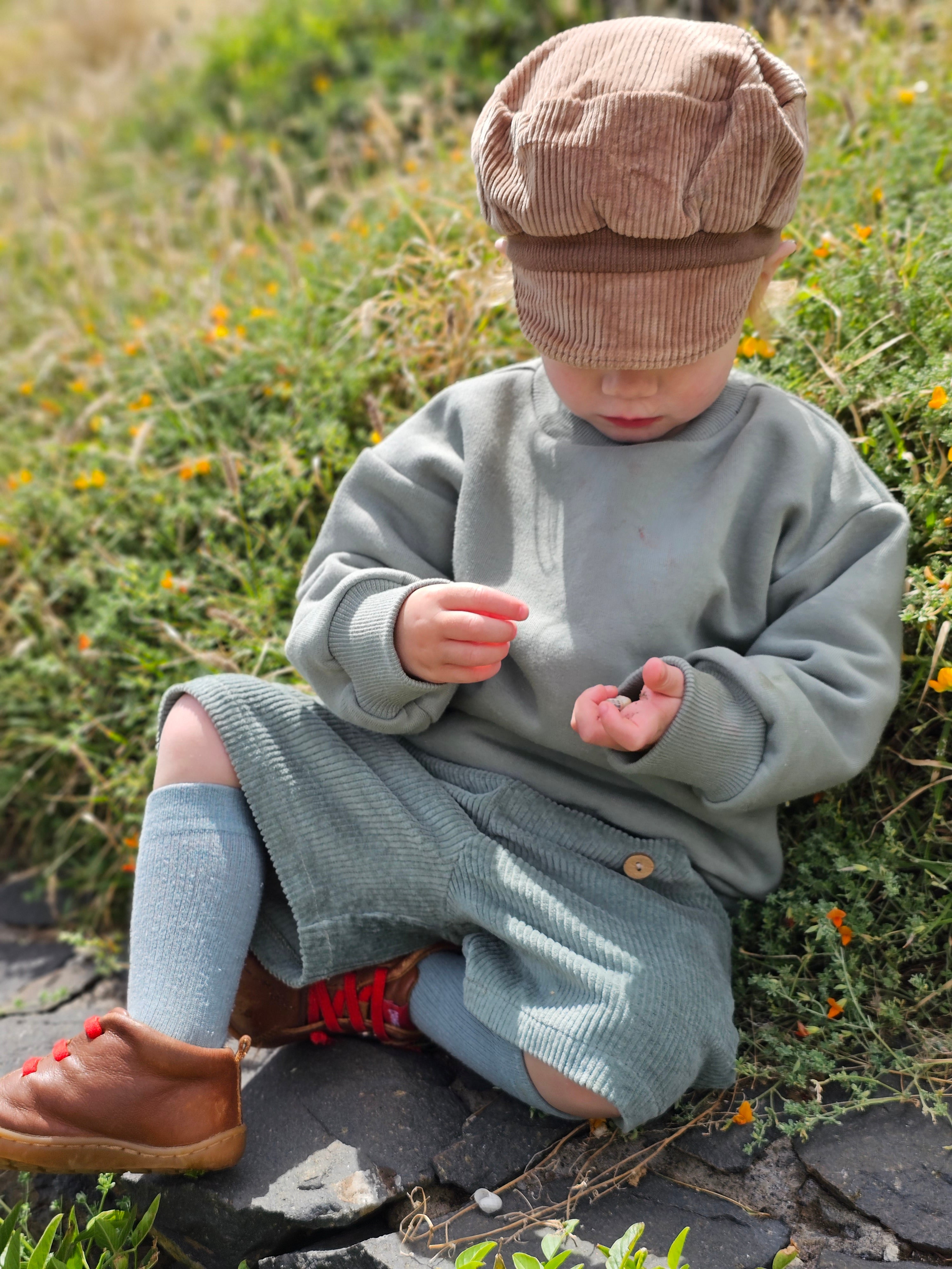 Ein Kind trägt die Kurze Buxe Breitcord - Aqua, sitzt auf einem Stein, umgeben von Gras und Blüten, und inspiziert ein kleines Objekt. Cordhose robust und perfekt für Abenteuer.