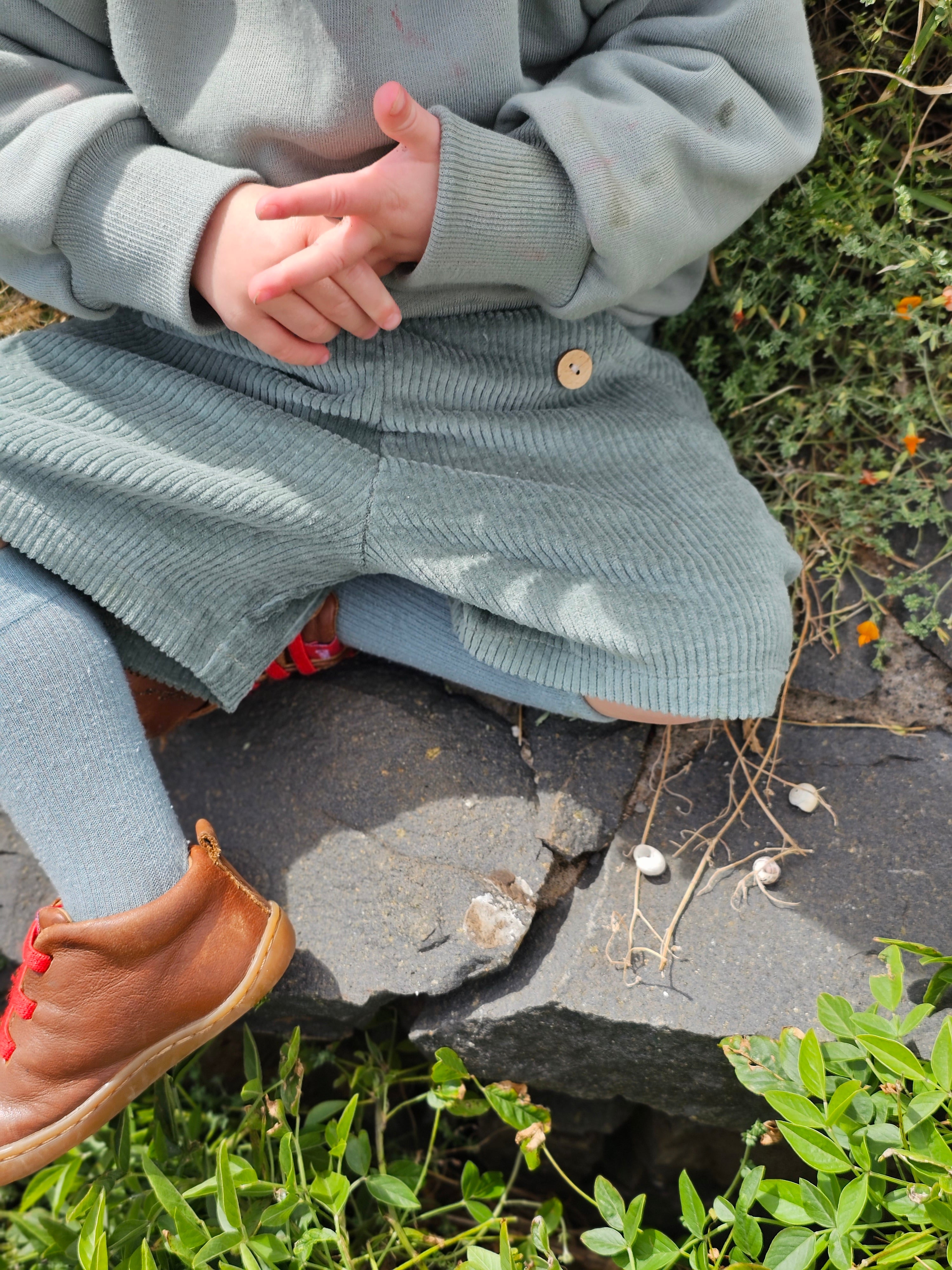 Kind trägt Kurze Buxe Breitcord - Aqua mit blauem Unterton, über blauen Strumpfhosen. Die Hose ist locker geschnitten, aus robustem Cord, ideal für aktive Abenteuer. Hände im Schoß gefaltet.