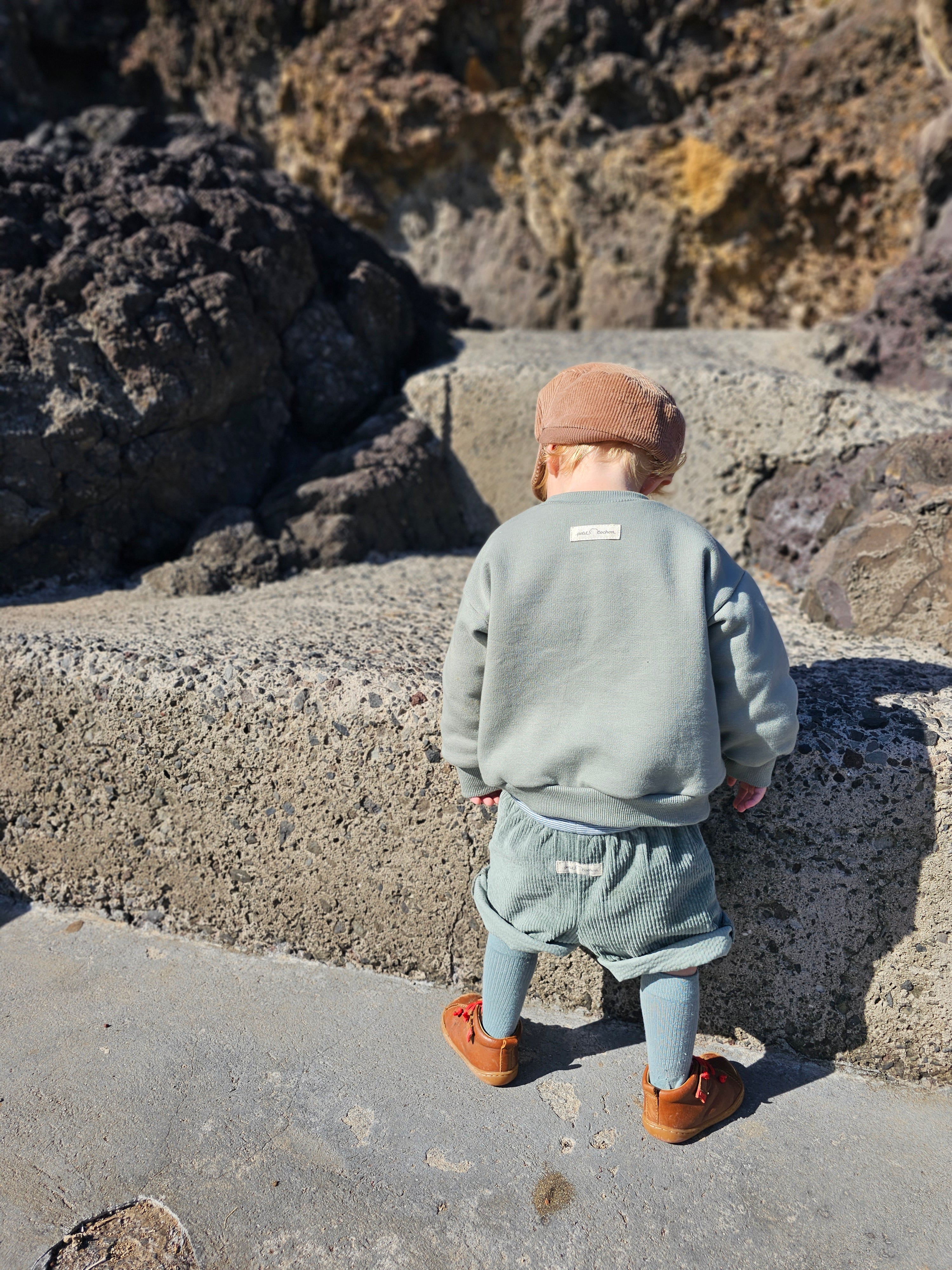 Ein Kind trägt die Kurze Buxe Breitcord - Aqua, steht auf einer gepflasterten Fläche neben einer niedrigen Steinmauer. Die Hose ist aus weichem Breitcord, perfekt für Abenteuer.