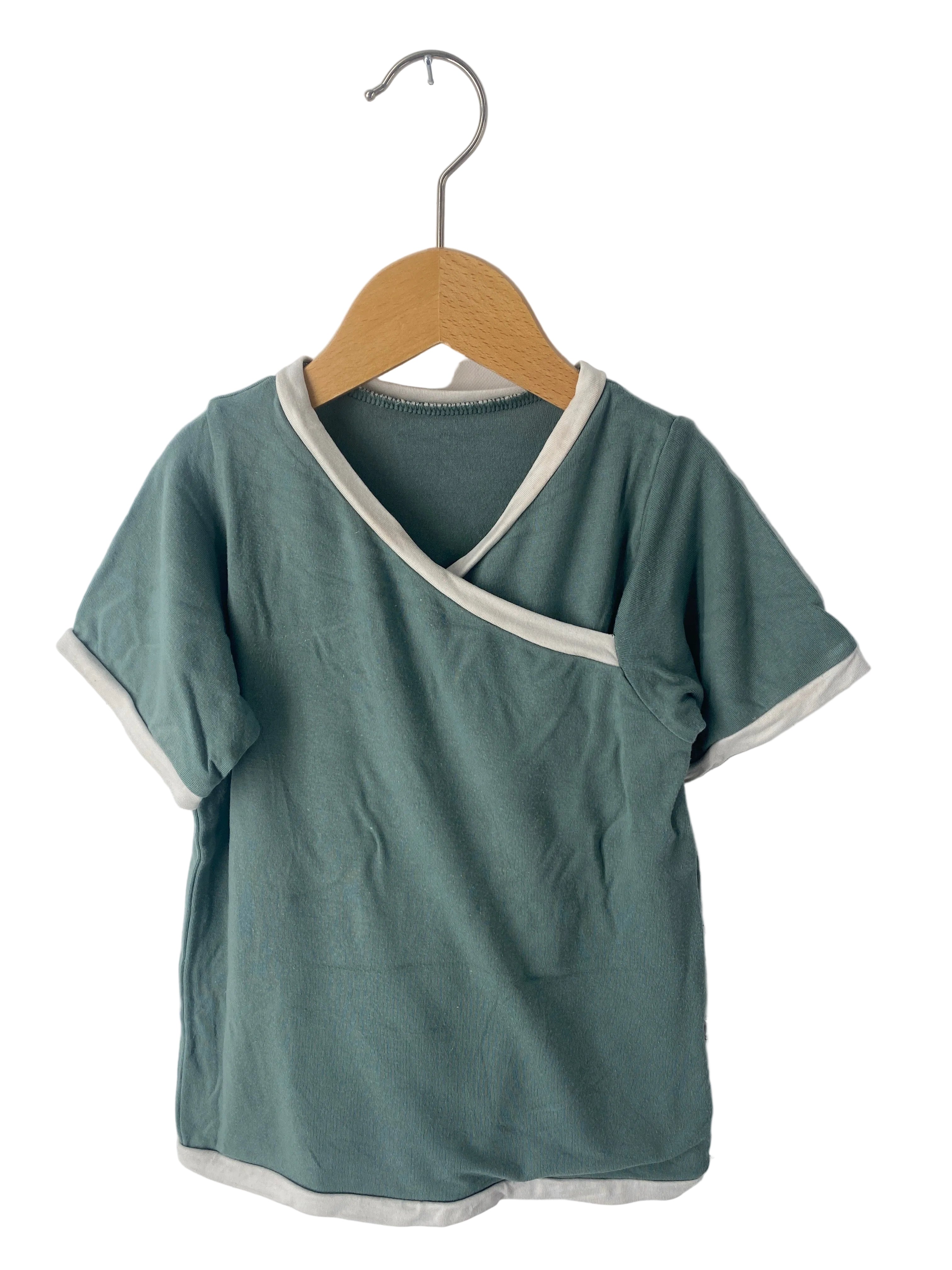 Ringer Shirt Bambusviskose Jade Gr 3Y, ein kurzärmeliges Wickelshirt für Kinder, in gutem Zustand mit leichten Flecken. Es hängt an einem Holzkleiderbügel, weißer Saum an V-Ausschnitt und Ärmeln.