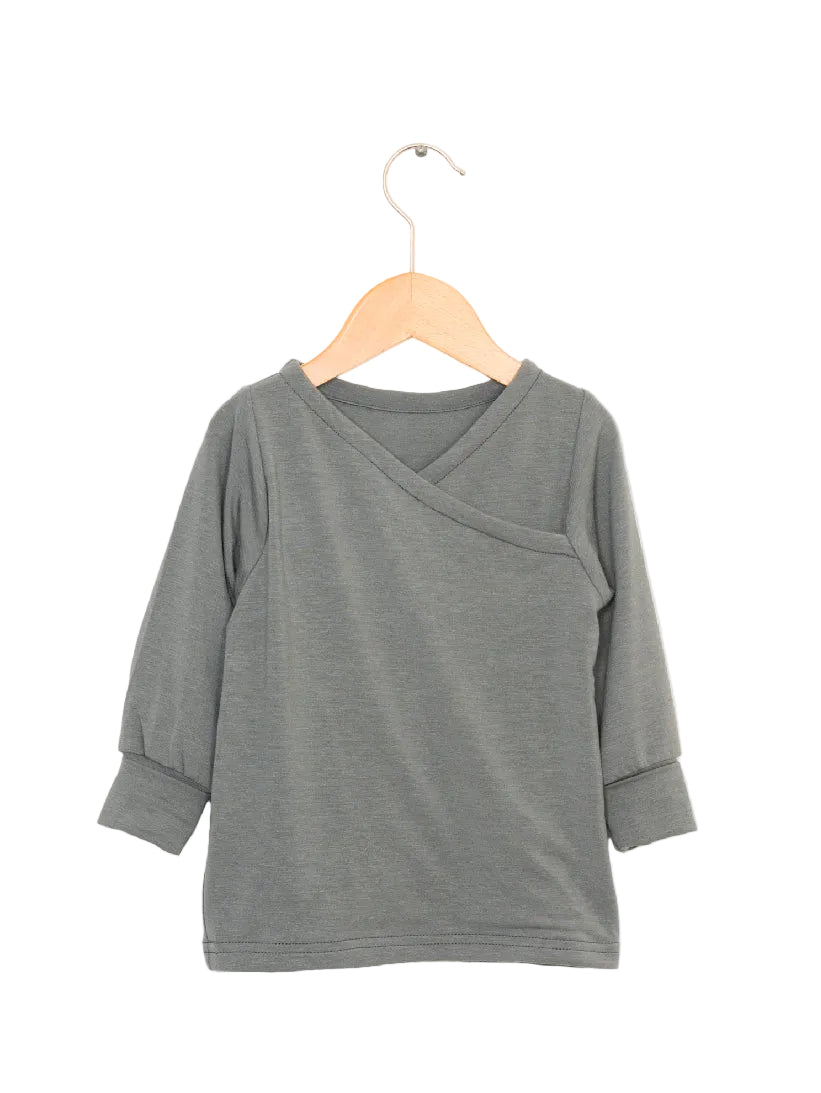 Wickelshirt Kaschmir Mix Taube Gr 1Y: Kinder-Langarmshirt mit Wickelausschnitt, Bündchen an den Ärmeln und geradem Saum, präsentiert auf einem Holzkleiderbügel vor weißem Hintergrund.