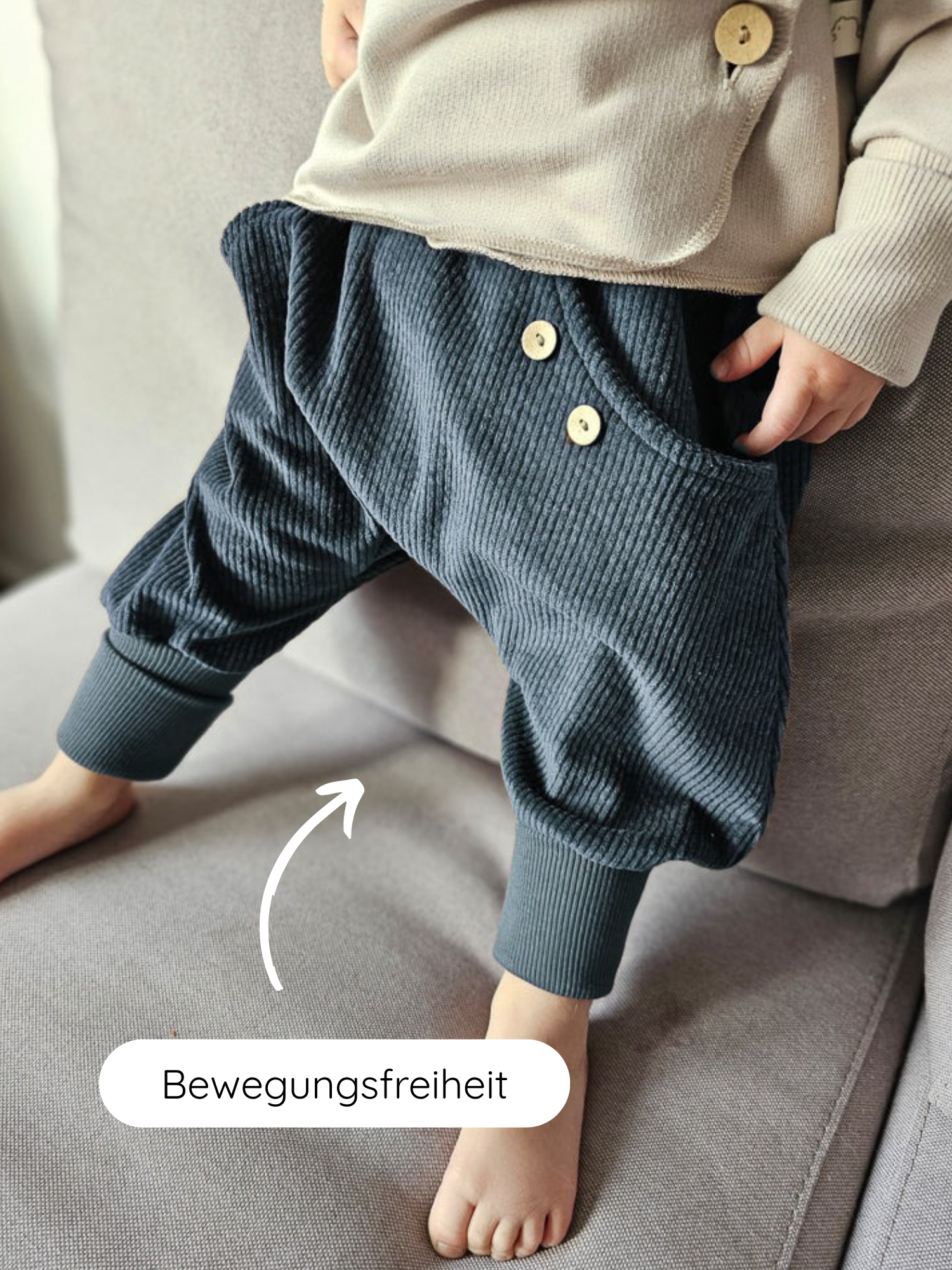Kind trägt Knickerbocker Breitcord - Jeansblau auf grauem Sofa, mit weitem Schrittbereich und Holzköpfen, ideal für Bewegungsfreiheit. Kombiniert mit beigem Oberteil, illustriert durch weißen Pfeil und „Bewegungsfreiheit“.