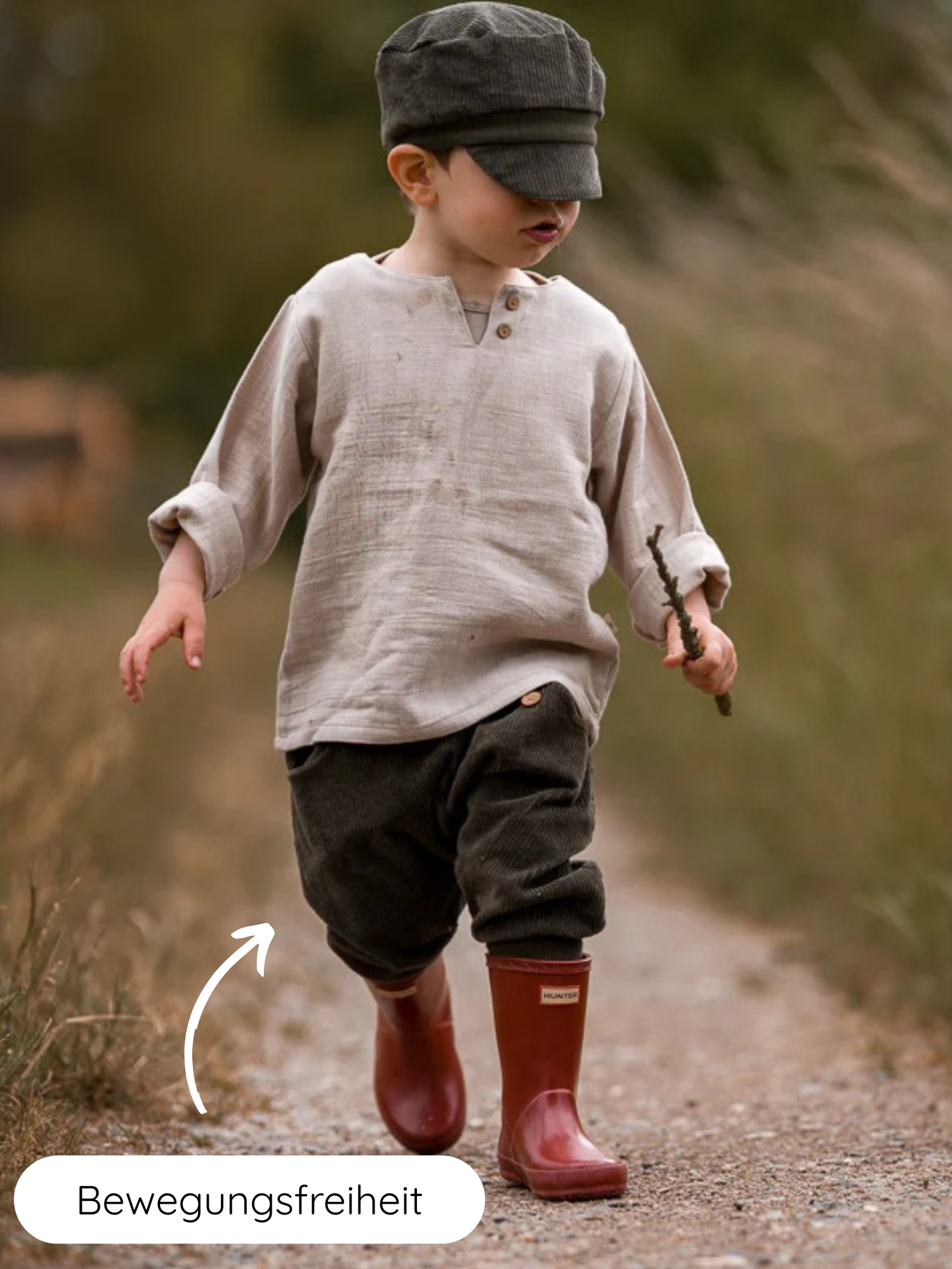 Ein kleiner Junge trägt die Knickerbocker Breitcord - Kiwi, hervorgehoben durch Bewegungsfreiheitspfeil, mit lockerem Schnitt und roten Gummistiefeln, ideal für aktive Kinder.