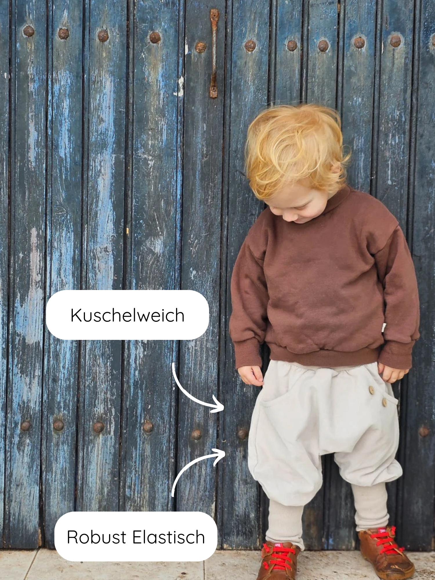 Kleinkind in braunem Sweatshirt und Knickerbocker Feincord - Sand vor blauer Holztür. Die Hose ist kuschelweich und robust elastisch, ideal für aktive Kinder.