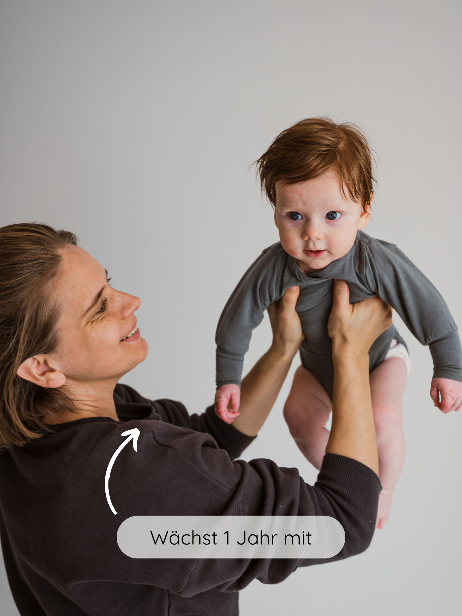 Junge Frau hebt Baby im Langarm-Loopbody Kaschmir-Mix - Taube lächelnd hoch. Der Body ist weich, ohne Knöpfe, und wächst ein Jahr mit, ideal für zarte Babyhaut.