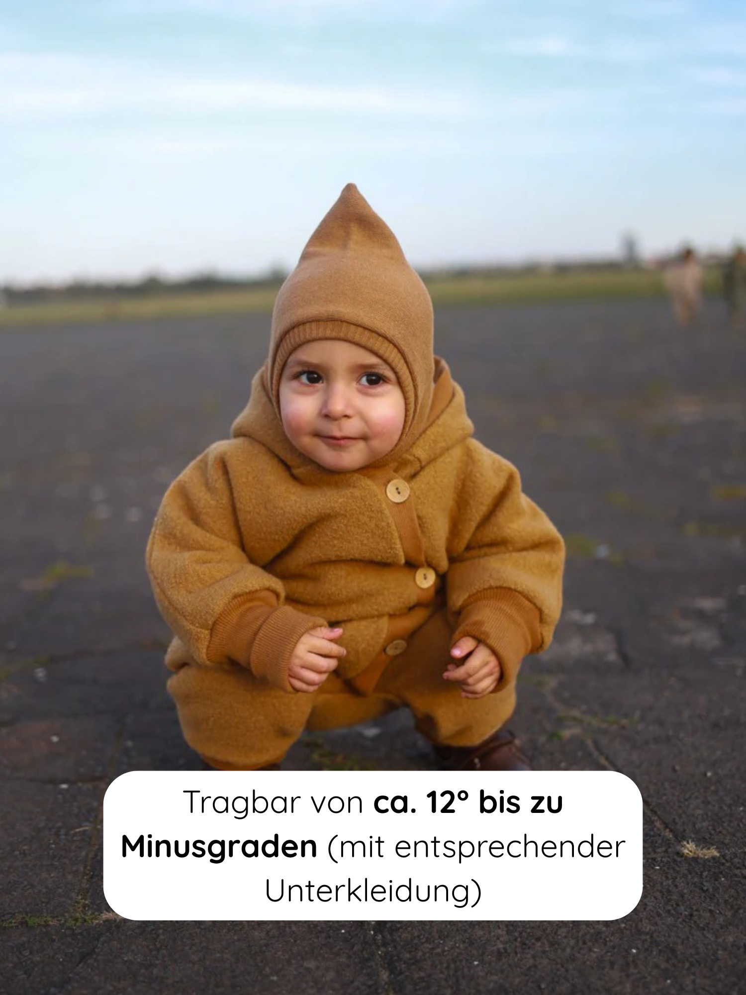 Kleinkind in einem Winter Wollwalk Overall mit Teddyfutter - Honig, kniend auf Asphalt. Der Overall hat eine spitze Kapuze, breite Bündchen und bietet Platz für Schichten darunter. Ideal für kaltes Wetter.
