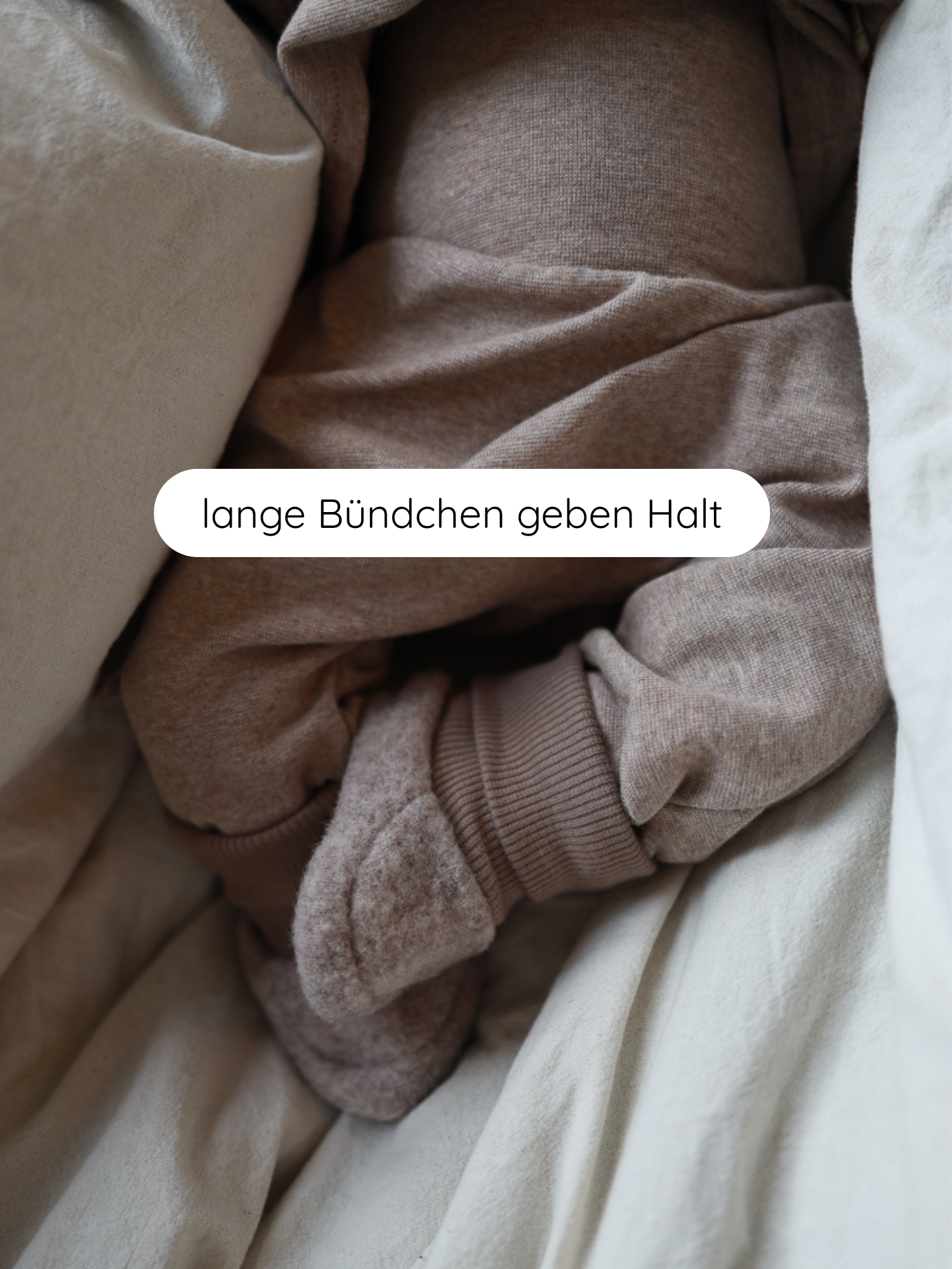 Babybooties Merino Wollfleece - Hafer: Nahaufnahme eines sanft strukturierten, hellbraunen Babystramplers mit extra langen, gerippten Bündchen, umgeben von neutraler Bettwäsche. Der Satz lange Bündchen geben Halt ist überlagert.