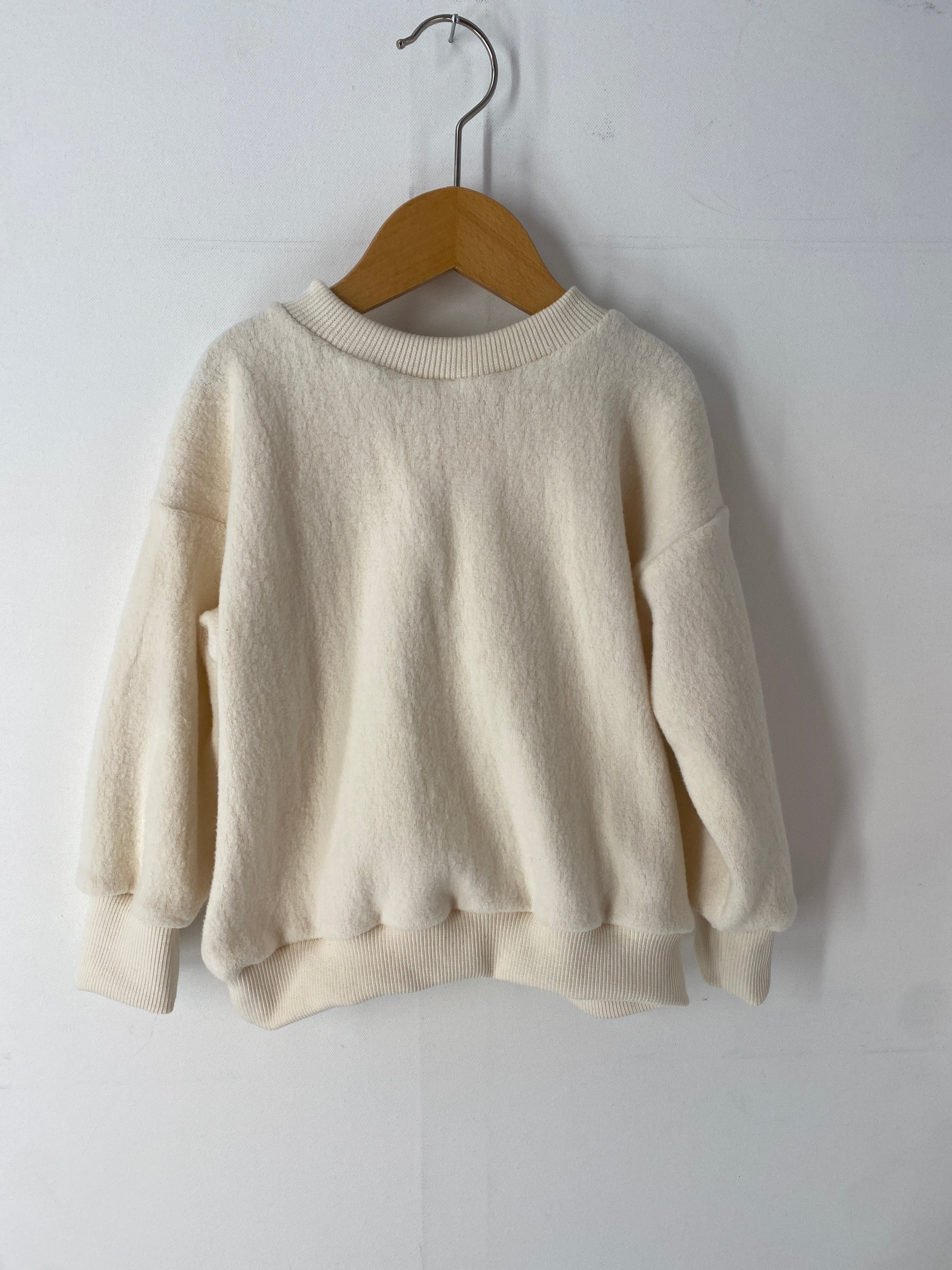 NEU Pulli aus Bio-Teddy Gr 1Y hängt an einem Holzbügel. Der weiße, gestrickte Pullover zeigt eine weiche Textur, ideal für einjähriges Kind, präsentiert auf einem schlichten Holzrahmen.
