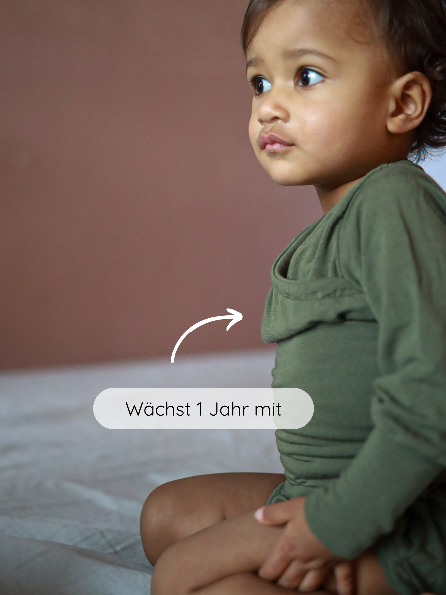Ein Kleinkind sitzt auf einem Bett und trägt den Loopbody Kaschmir-Mix - Pistazie, ein olivgrüner Langarm-Romper mit überlappender Front, der mit dem Kind ein Jahr mitwächst.