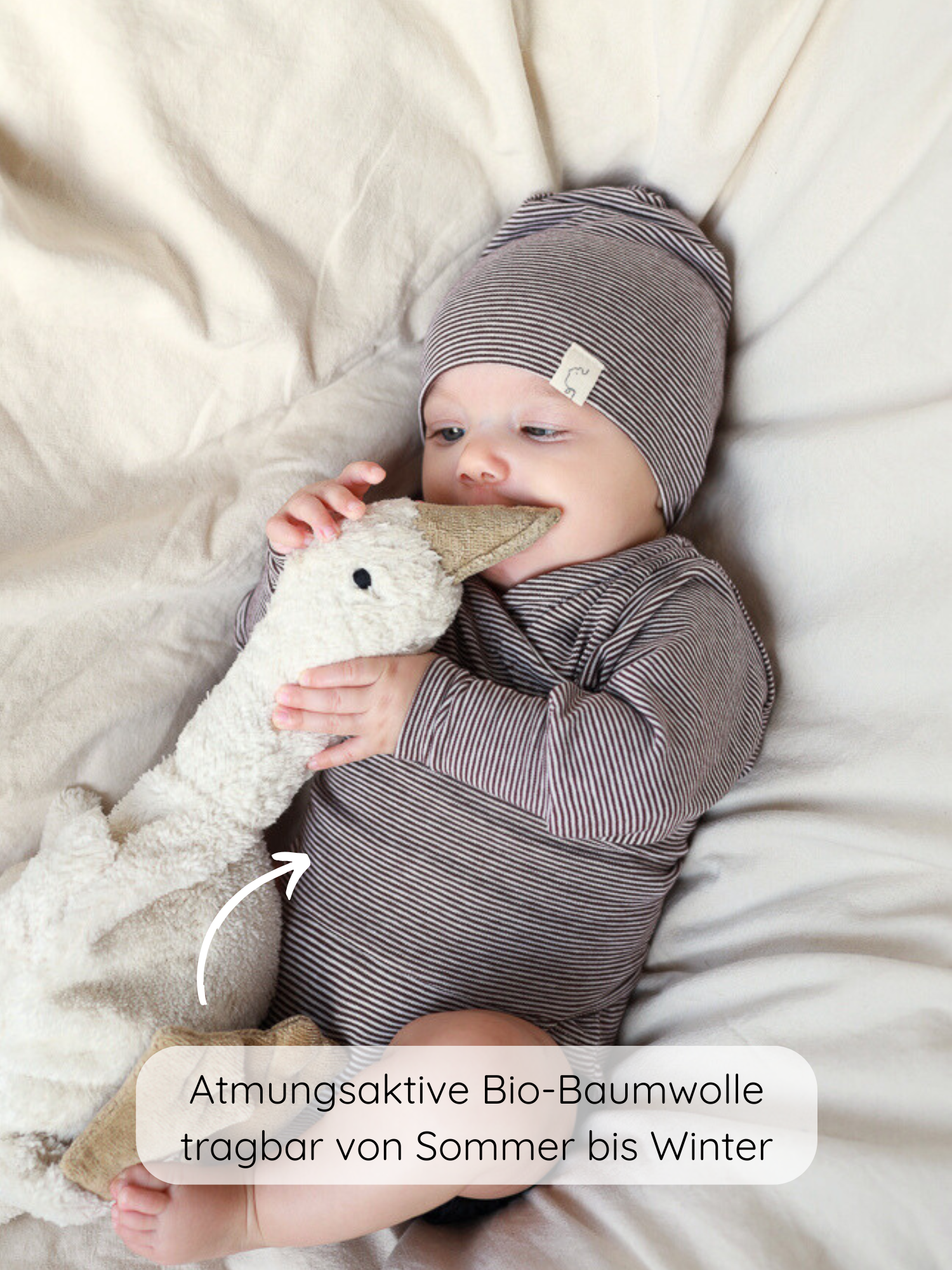 Ein Baby trägt den Loopbody Bio-Baumwolle-Ringel Schoko, bestehend aus einem gestreiften langärmeligen Body und einer Mütze, während es ein Plüschgans-Spielzeug hält. Kleidung aus atmungsaktiver Bio-Baumwolle.