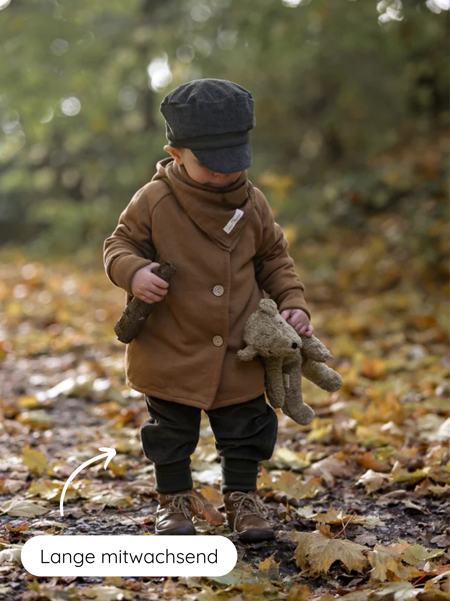Ein Kleinkind steht im Herbstlaub, trägt Knickerbocker Breitcord - Kiwi, braunen Mantel, dunkle Mütze, hält einen Stock und Teddy. Pfeil zeigt auf anpassbare Hosenbeine mit Lange mitwachsend.