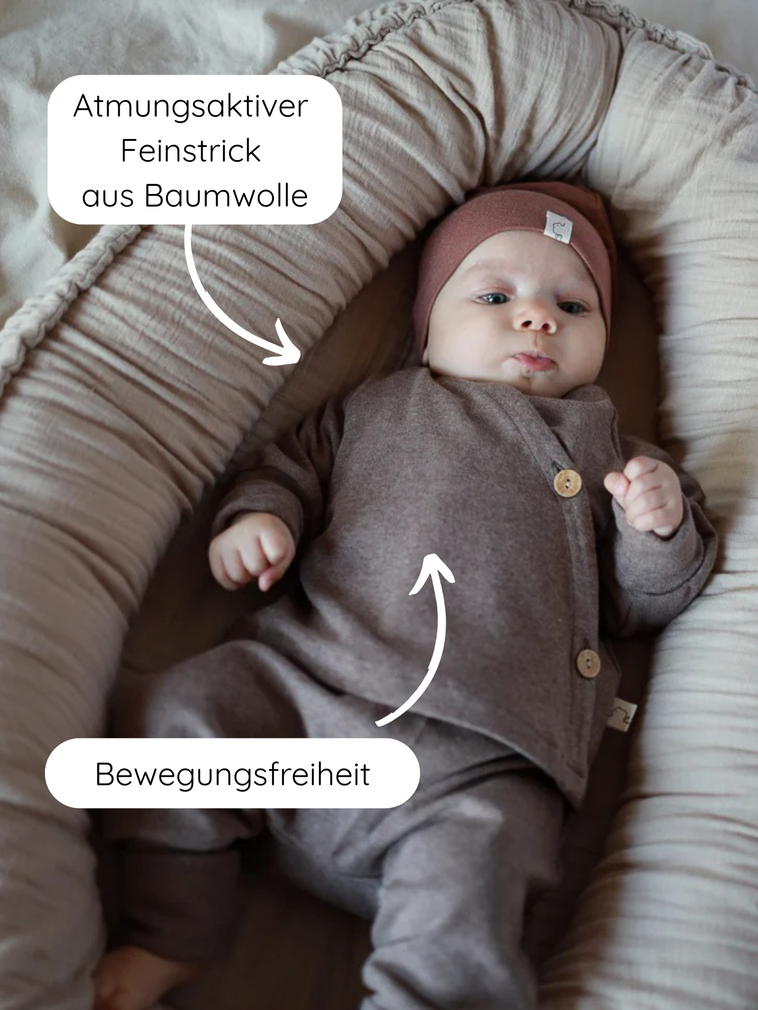 Baby in einem „Nest“-Bettchen, trägt Babyschlüttli Feinstrick - Praline Meliert, bestehend aus Jacke mit Holzknöpfen, Hose, Mütze; atmungsaktiver, elastischer Stoff bietet Bewegungsfreiheit.