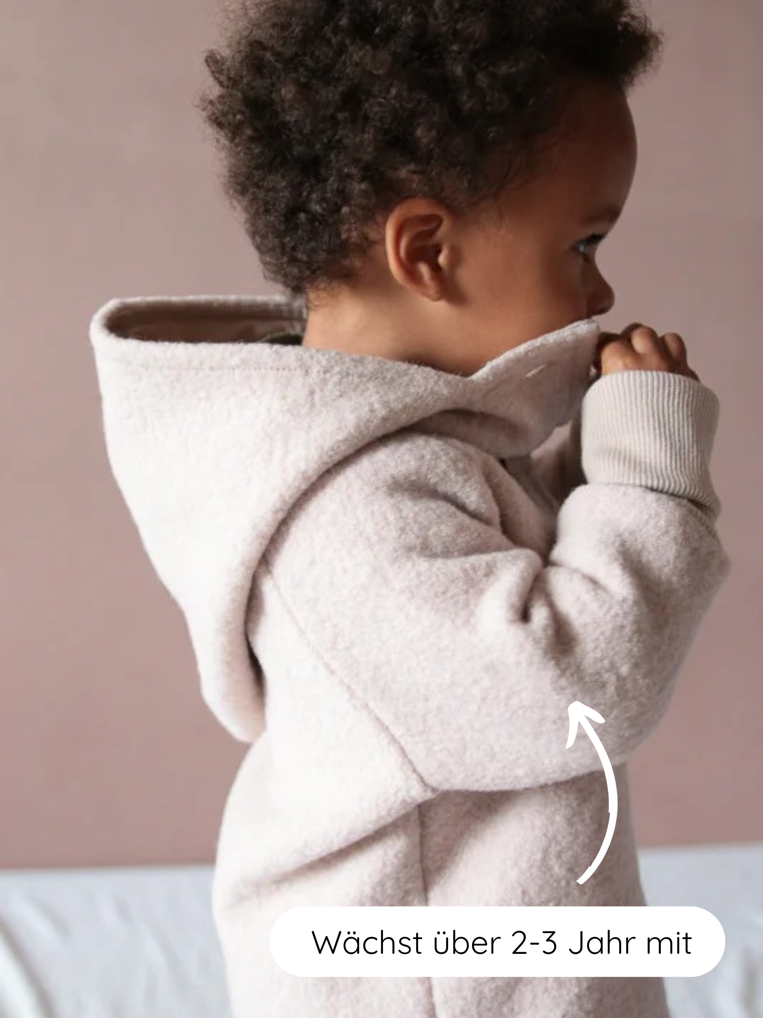 Ein Kleinkind im Profil trägt die Wichteljacke Wollwalk - Sand, eine beige, fleeceartige Kapuzenjacke. Ein Pfeil zeigt auf den Ärmel mit dem Text „Wächst über 2-3 Jahr mit“.