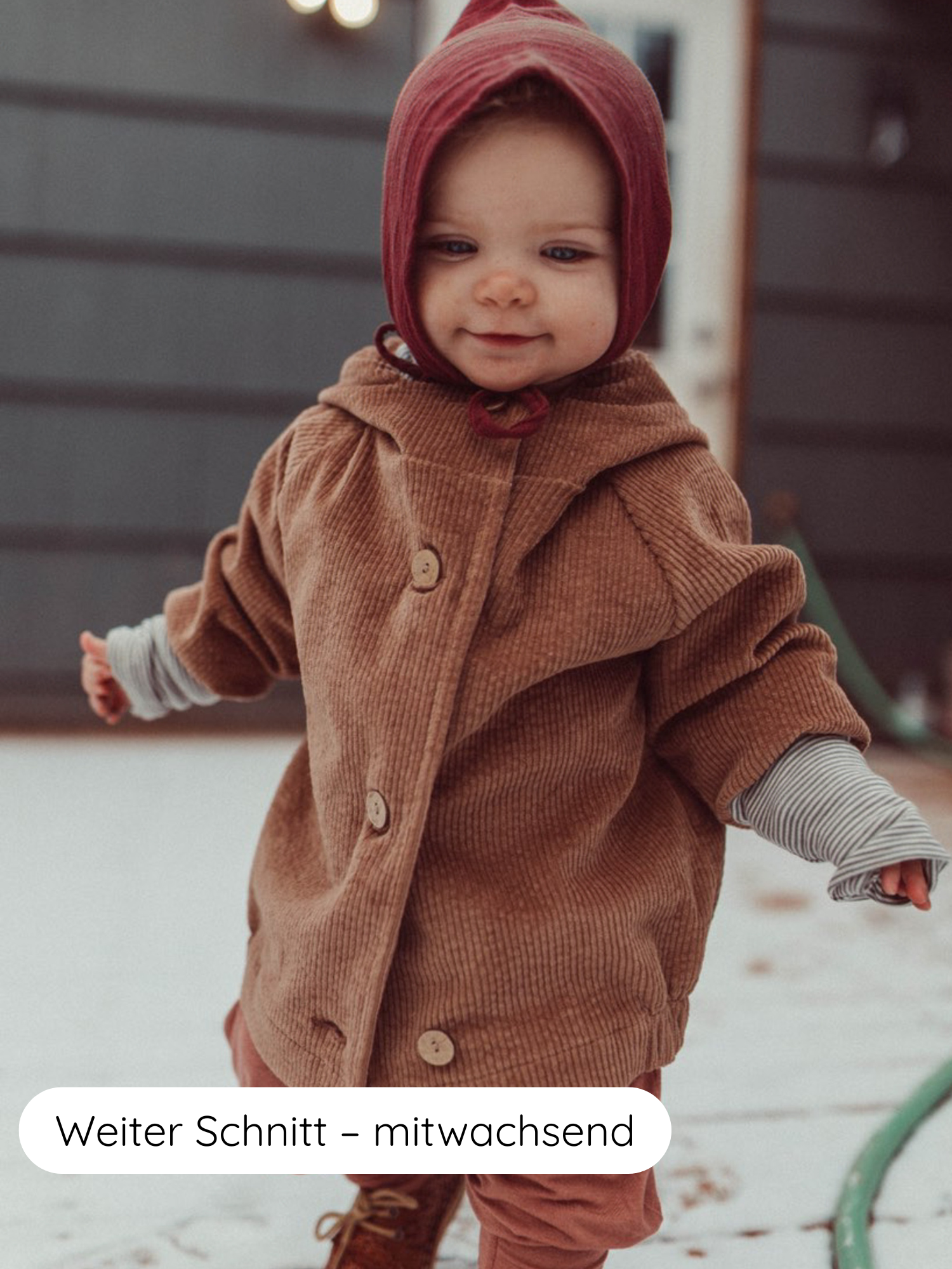Ein Kleinkind trägt die Wichteljacke Breitcord Stretch - Weizen, eine rust-braune Cordjacke mit großen Knöpfen und sichtbaren gestreiften Armbündchen, auf einem schneebedeckten Deck stehend.