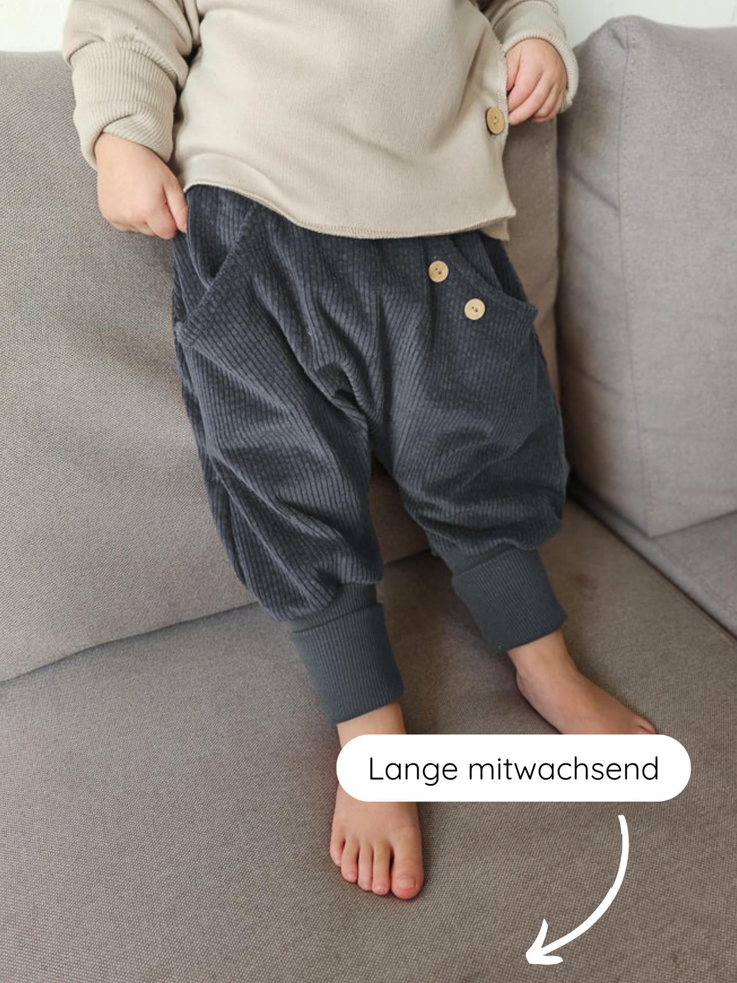 Kleinkind steht auf grauem Sofa, trägt Knickerbocker Breitcord - Jeansblau mit Knopfdetails und breiten Bündchen. Die Lange mitwachsend-Markierung zeigt, dass die Hose mit dem Kind mitwächst.