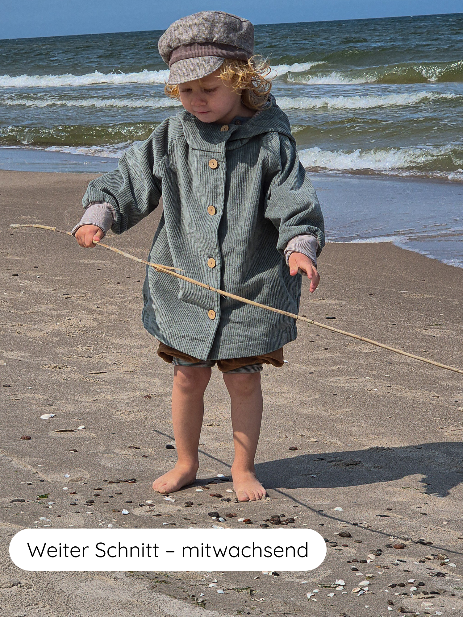 Ein Kind am Strand trägt die Wichteljacke Breitcord Stretch- Aqua, eine kapuzenartige Cordjacke mit krempelbaren Armbündchen und lockerem Schnitt, ideal für mitwachsendes Tragen, mit einem Stock im Sand.