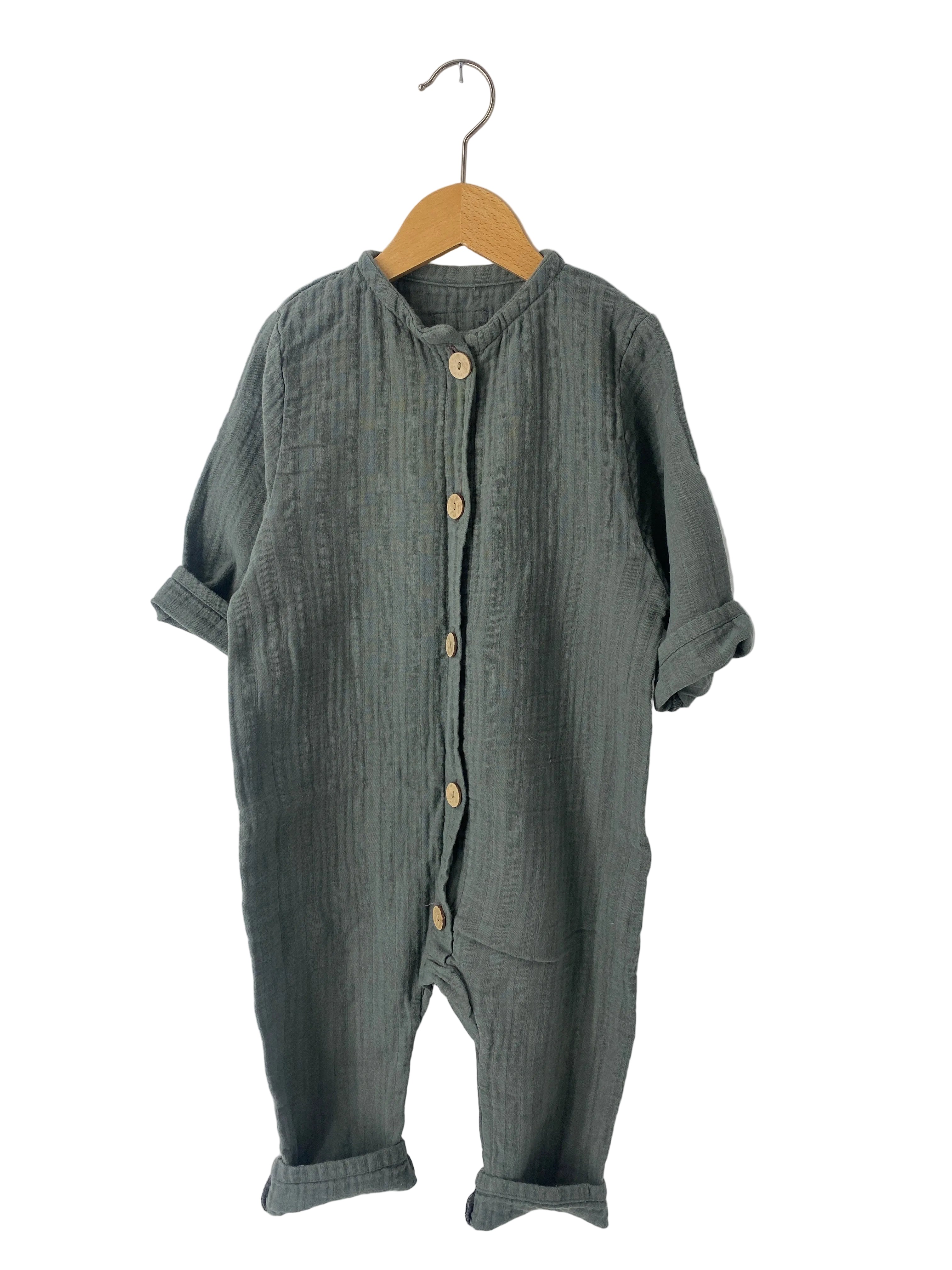 Mini Overall Musselin Taube Gr 2Y: Einteiler aus weichem, strukturiertem Stoff mit langen Ärmeln und Holzknöpfen. Der Zustand ist gut, jedoch mit leichten Flecken. Präsentiert auf einem Holzbügel vor weißem Hintergrund.