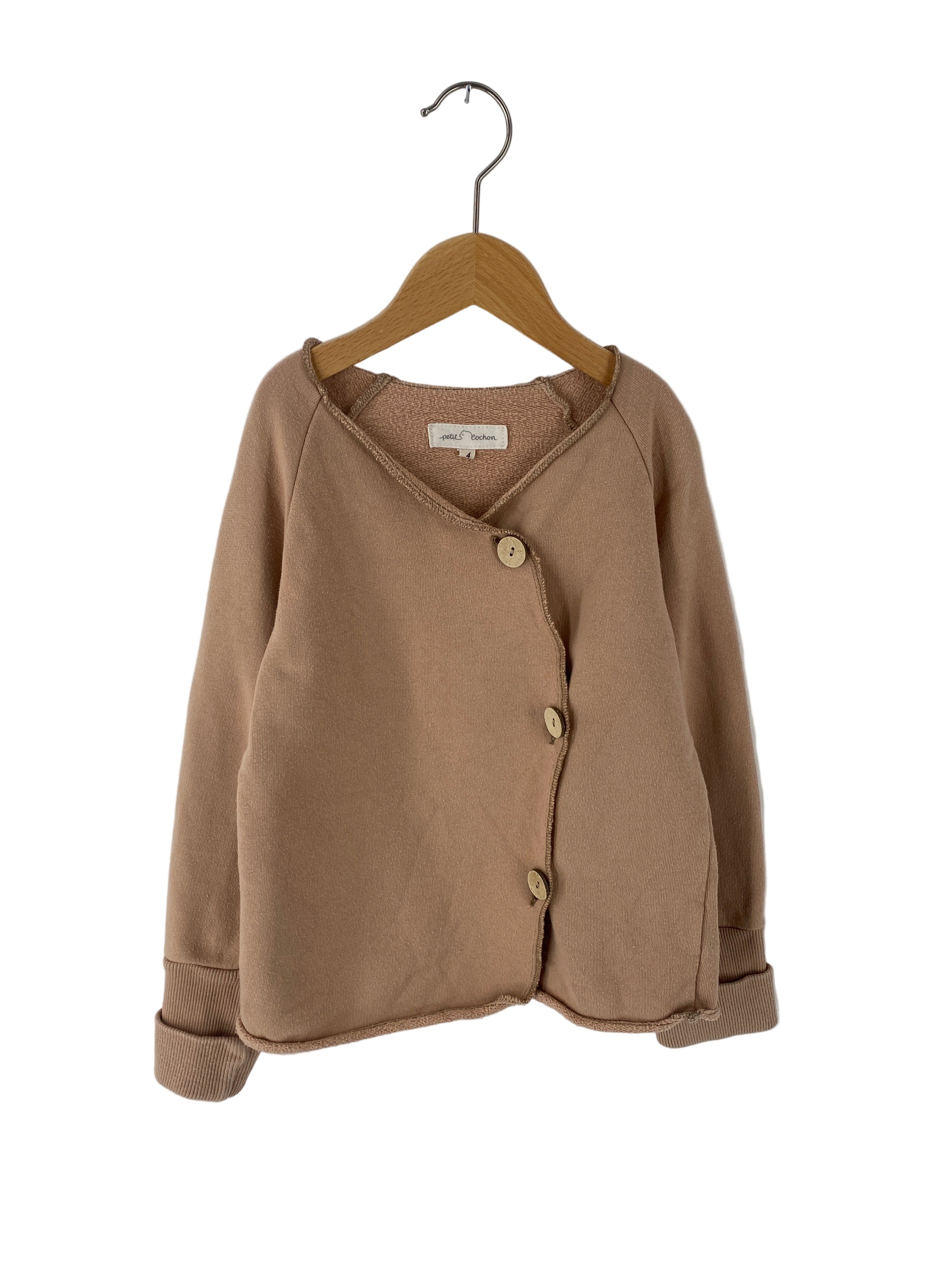 Schlüttli Bio-Sweat altes Puder Gr 4/5Y: Kindercardigan in hellem Braun mit langen Ärmeln, sichtbaren Nähten, drei seitlich versetzten Holzknöpfen und umgeschlagenen Bündchen. Hängt an einem Holzbügel, Label petit Cochon sichtbar.