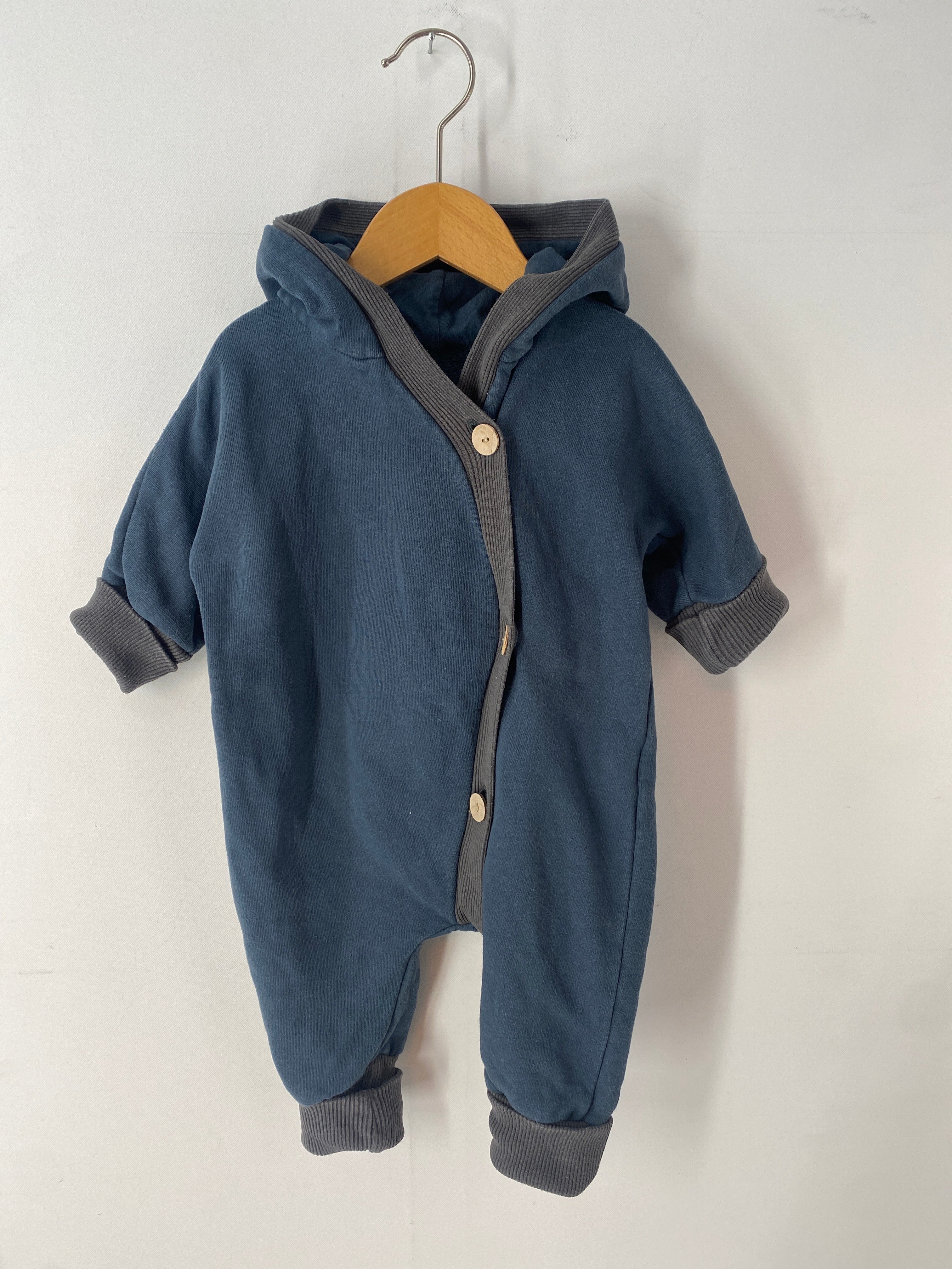 Kuschelanzug Bio-Sweat Marine Gr 3M auf einer Schaukel. Der blaue Babystrampler zeigt gestrickte Bündchen und eine Kapuze, ideal für Komfort und Wärme. Zustand: gut.