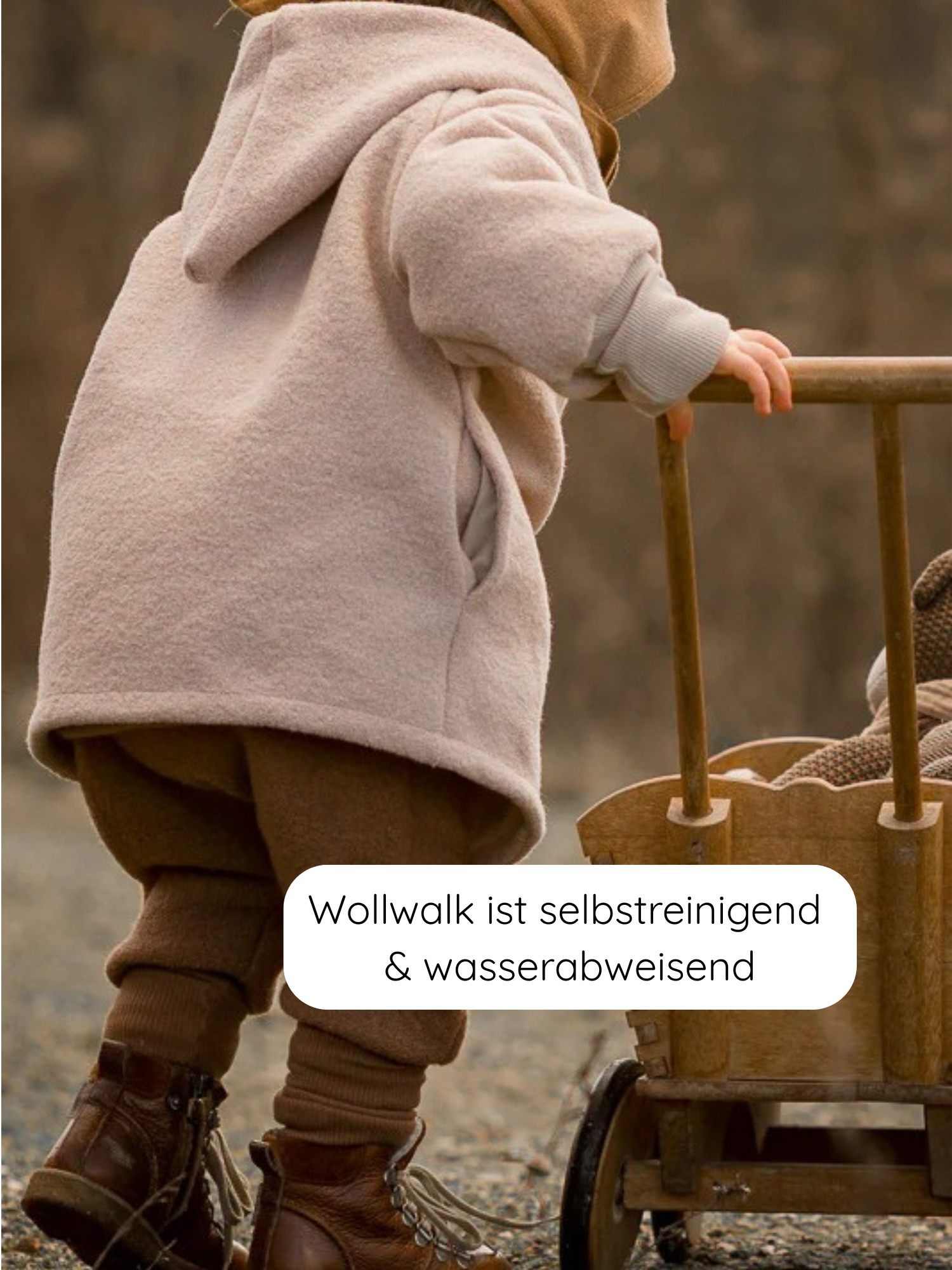 Kleinkind in Wichteljacke Wollwalk - Sand mit Bio-Jersey-Futter, braunen Wollhosen und dunkelbraunen Lederstiefeln, die Hand am Griff eines Holzspielwagens auf Kiesboden.