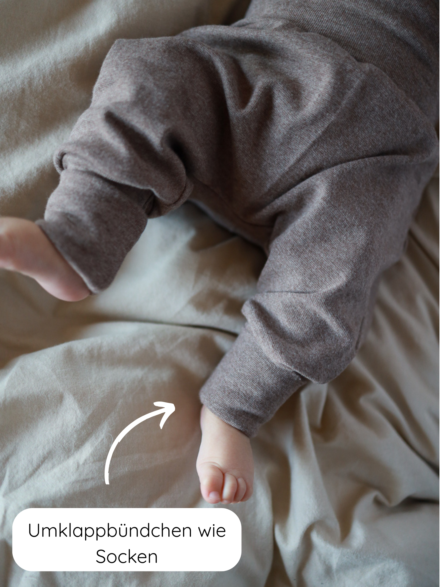 Nahaufnahme eines Babys in Babyknicker Feinstrick - Praline Meliert auf neutralem Untergrund. Die weichen, braunen Jerseyhosen haben umklappbare Bündchen, die wie Socken wirken, mit einem weißen Pfeil und deutscher Beschriftung.