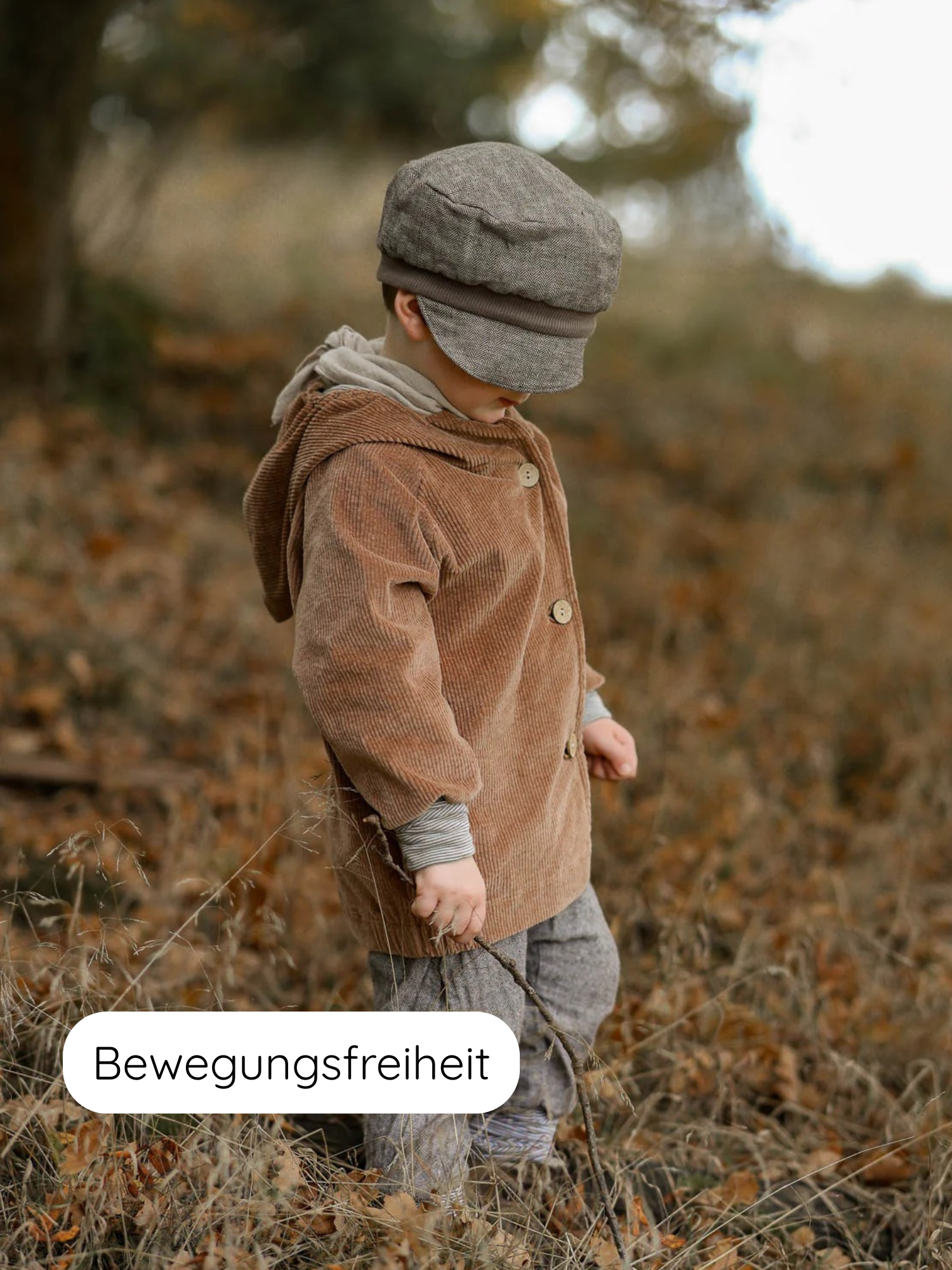 Kleinkind in Wichteljacke Breitcord Stretch - Weizen mit Wichtelkapuze und Ringel-Armbündchen, hält einen Zweig in einem herbstlichen Feld. Der Text Bewegungsfreiheit ist links unten eingeblendet.