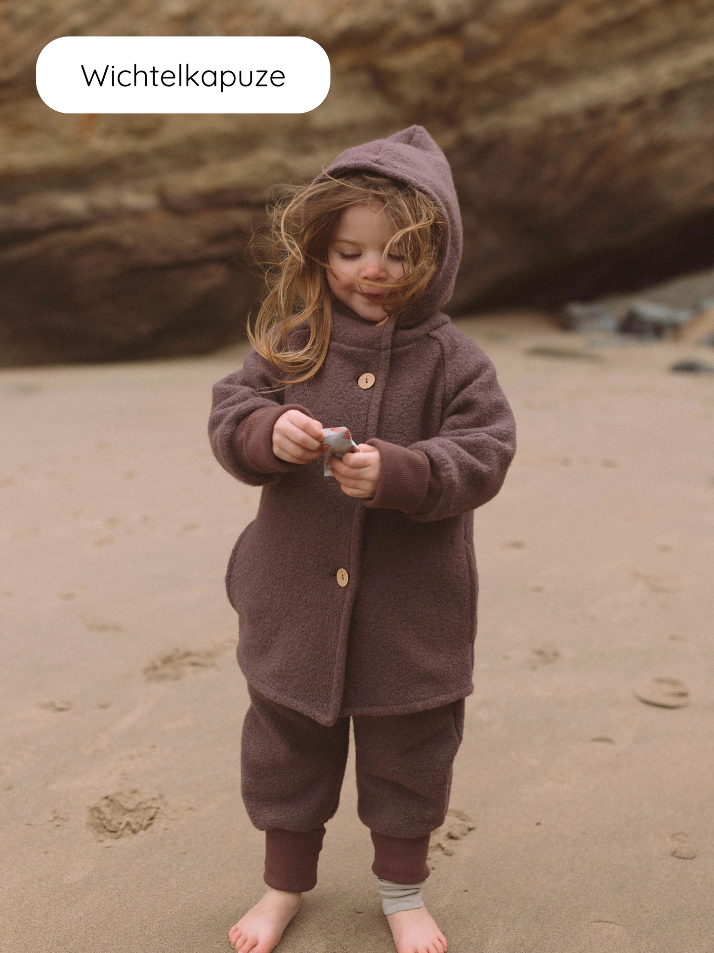 Kleinkind am Strand trägt die Wollwalk-Wichteljacke - Malve, mit spitzer Kapuze und krempelbaren Bündchen, hält einen kleinen Gegenstand. Perfekt für Herbst, Frühling und Winter, bietet Bewegungsfreiheit.